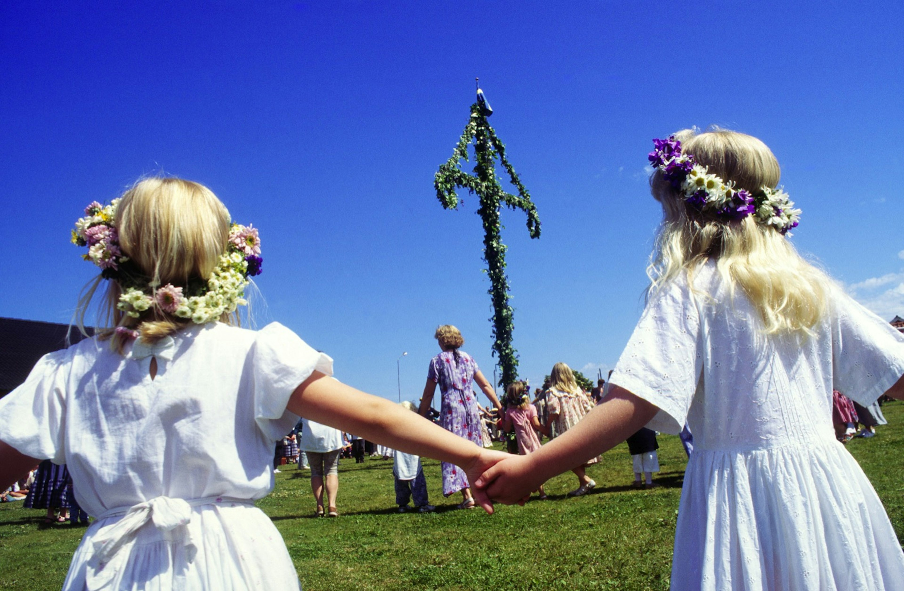 A pair of girls wearing white dresses and floral crowns hold hands as they dance around a maypole behind another group of people holding hands. Summer Solstice is known as Midsummer in Sweden.