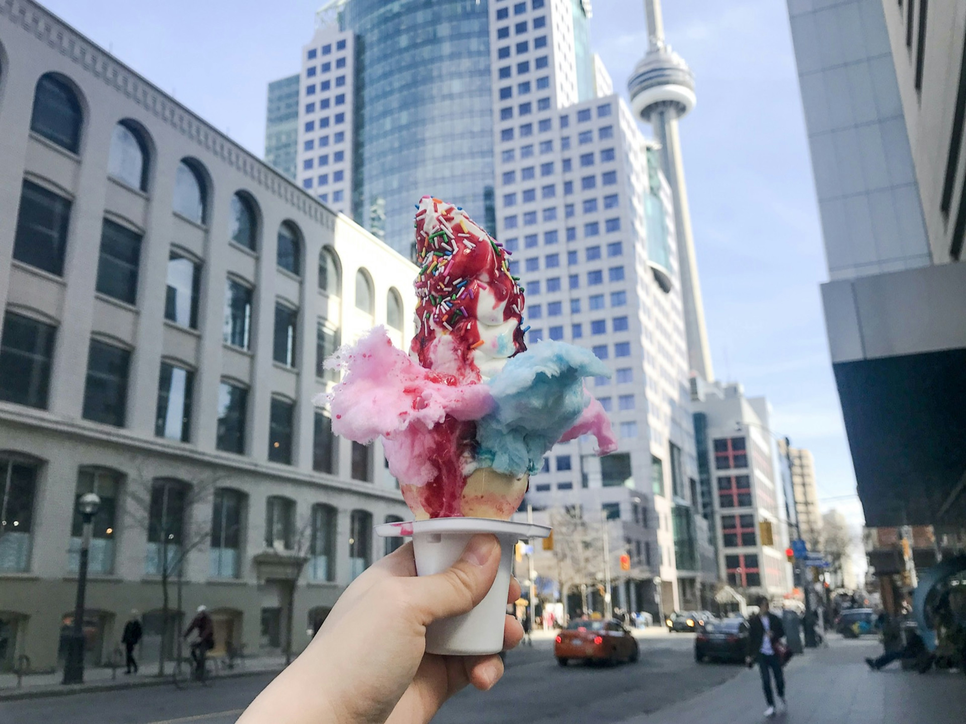 Tufts of pink and blue cotten candy poke out of an ice cream cone held in front of the CN tower in Toronto