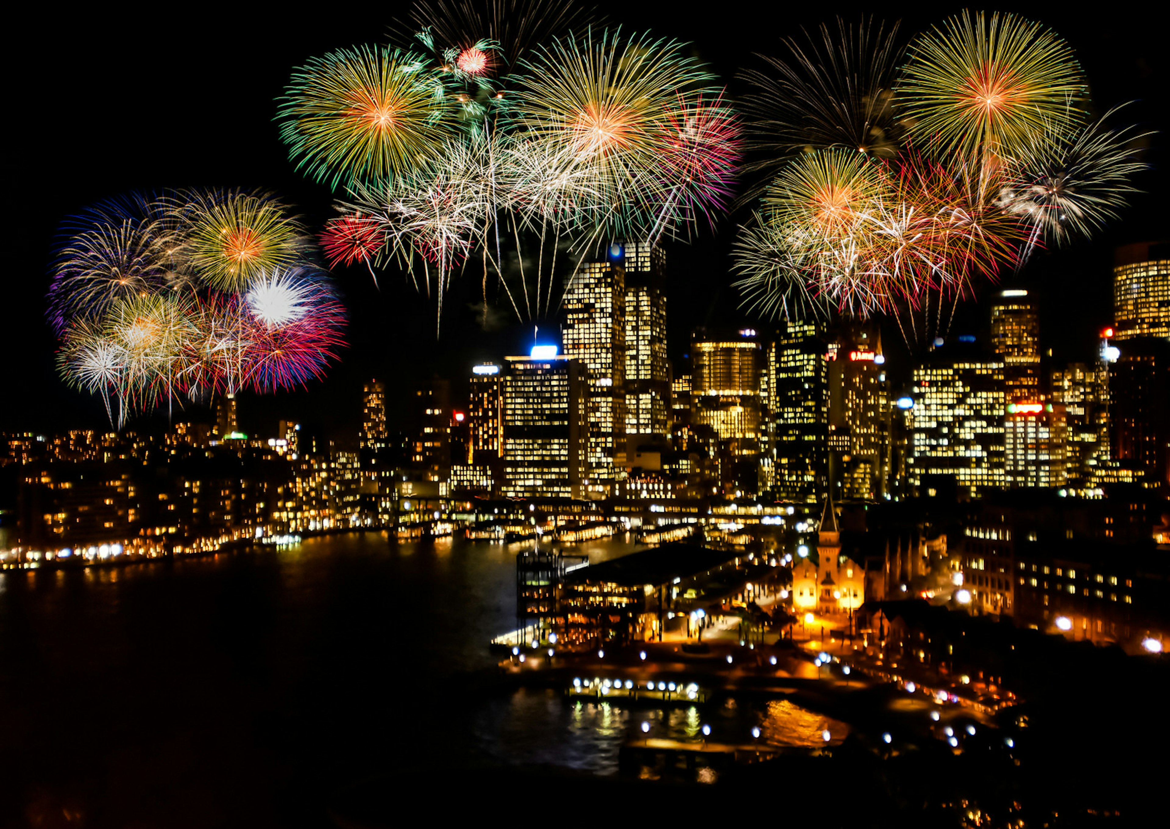 Sydney Harbour at nighttime with a colorful fireworks display happening over the water and skyscrapers