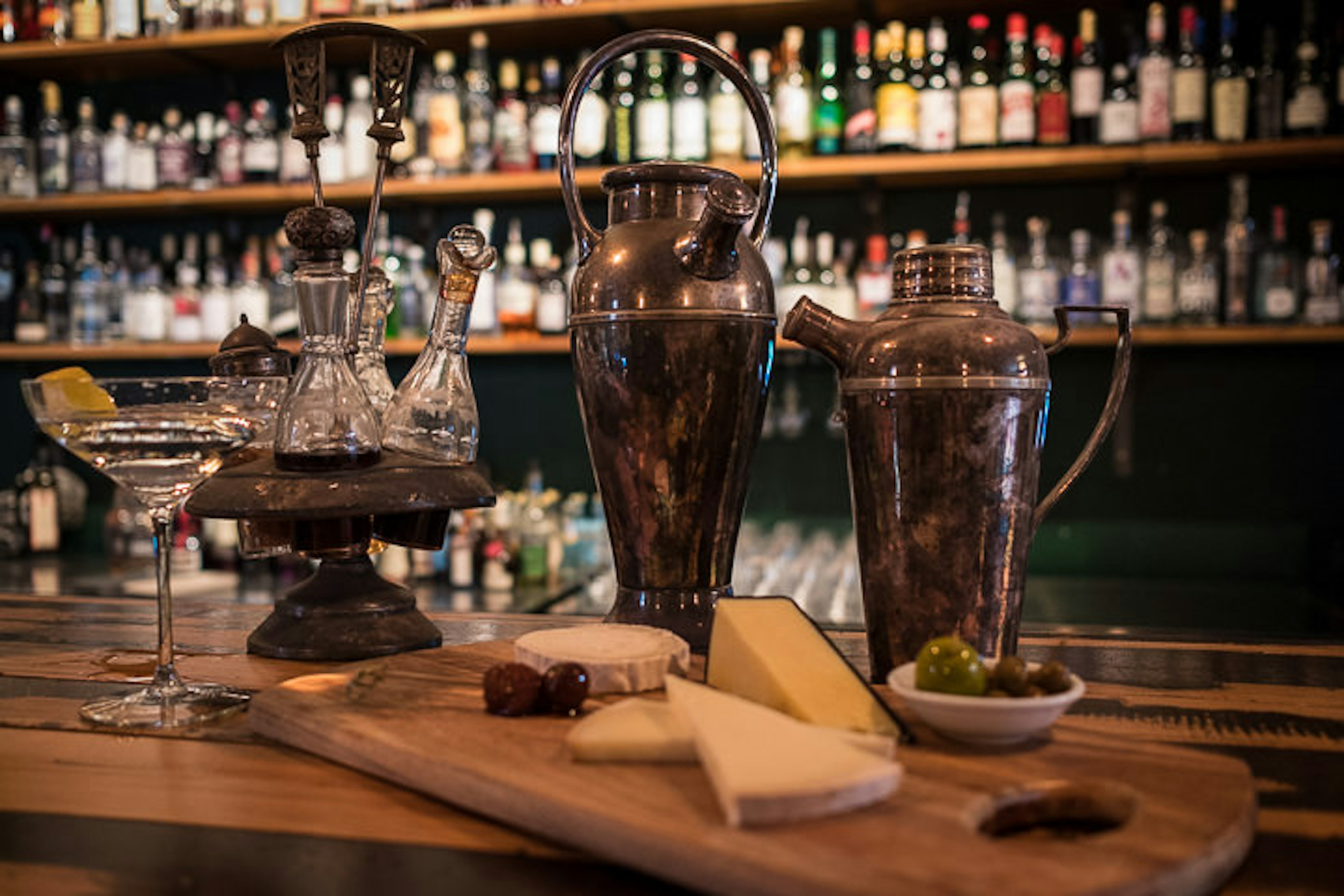 Vintage cocktail shakers sit on a wooden bar top alongside a wooden platter of cheese and olives as well as a martini glass. Out of focus in the background we can see shelves stacked with various gin bottles.