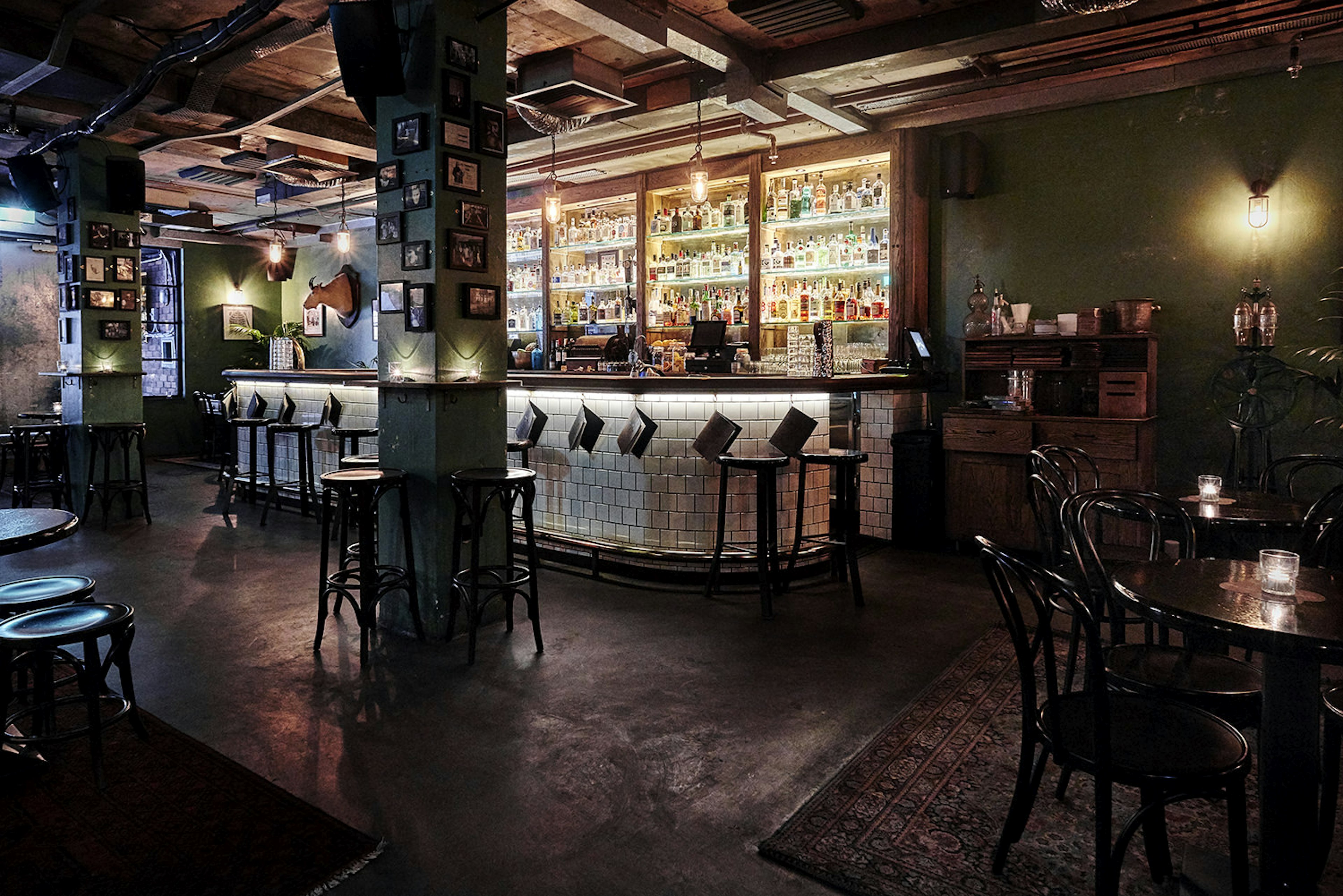 The dimly-lit bar at The Barber Shop, Sydney. The walls and pillars are hung with lots of small, framed photos. The bar is covered in white tiles and behind it there are illuminated glass shelves packed with bottles of gin. There are antique rugs on the ground and darkly-varnished wooden furniture.