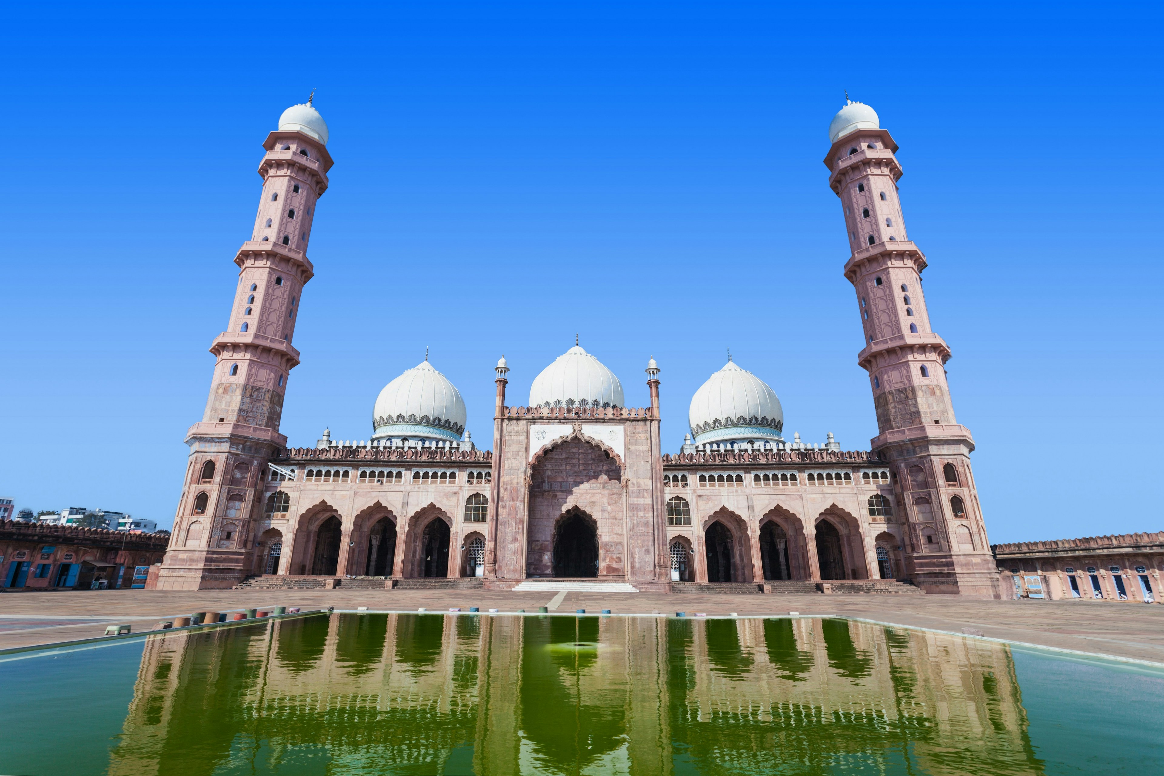 A view of Taj-ul-Masjid from the front, with the large mosque reflected in the pool used for washing in its central courtyard. The large mosque has three white domes and a towering minaret at both ends.