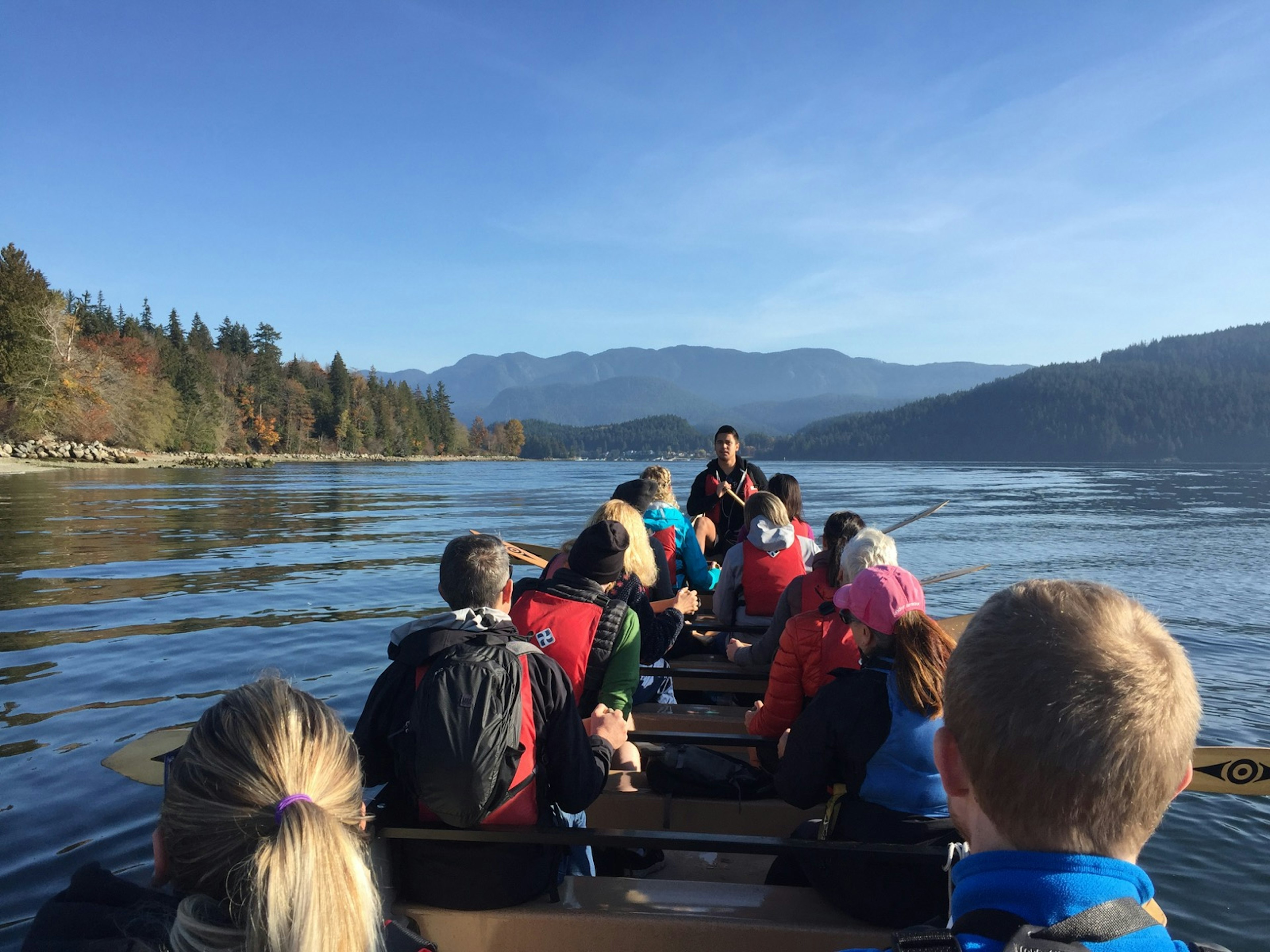 Looking forward in a canoe, several rows of paddlers propel a canoe through tranquil waters in British Columbia.