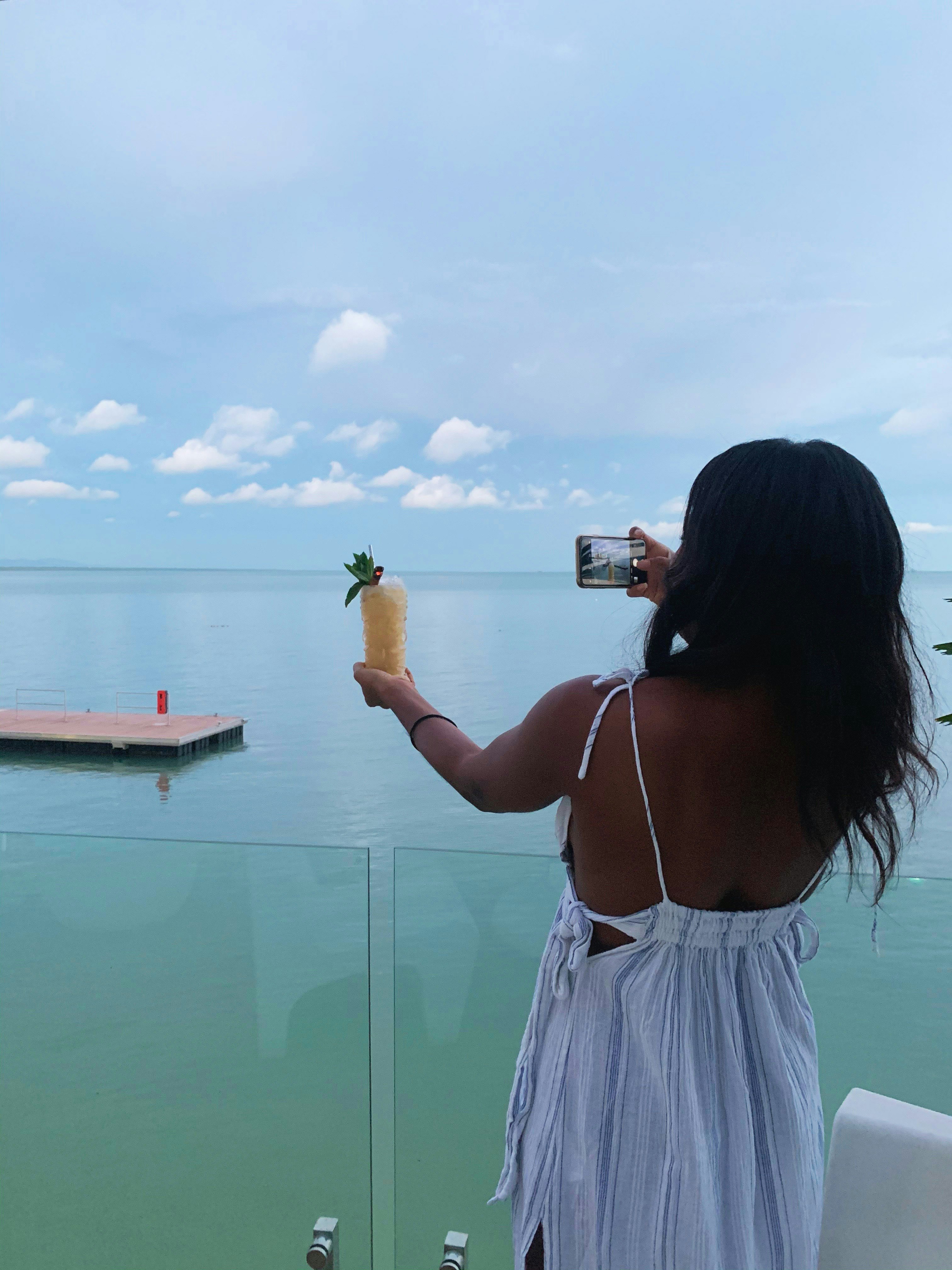 A woman holds up a drink with one hand while snapping a photo with her camera phone with her other hand.