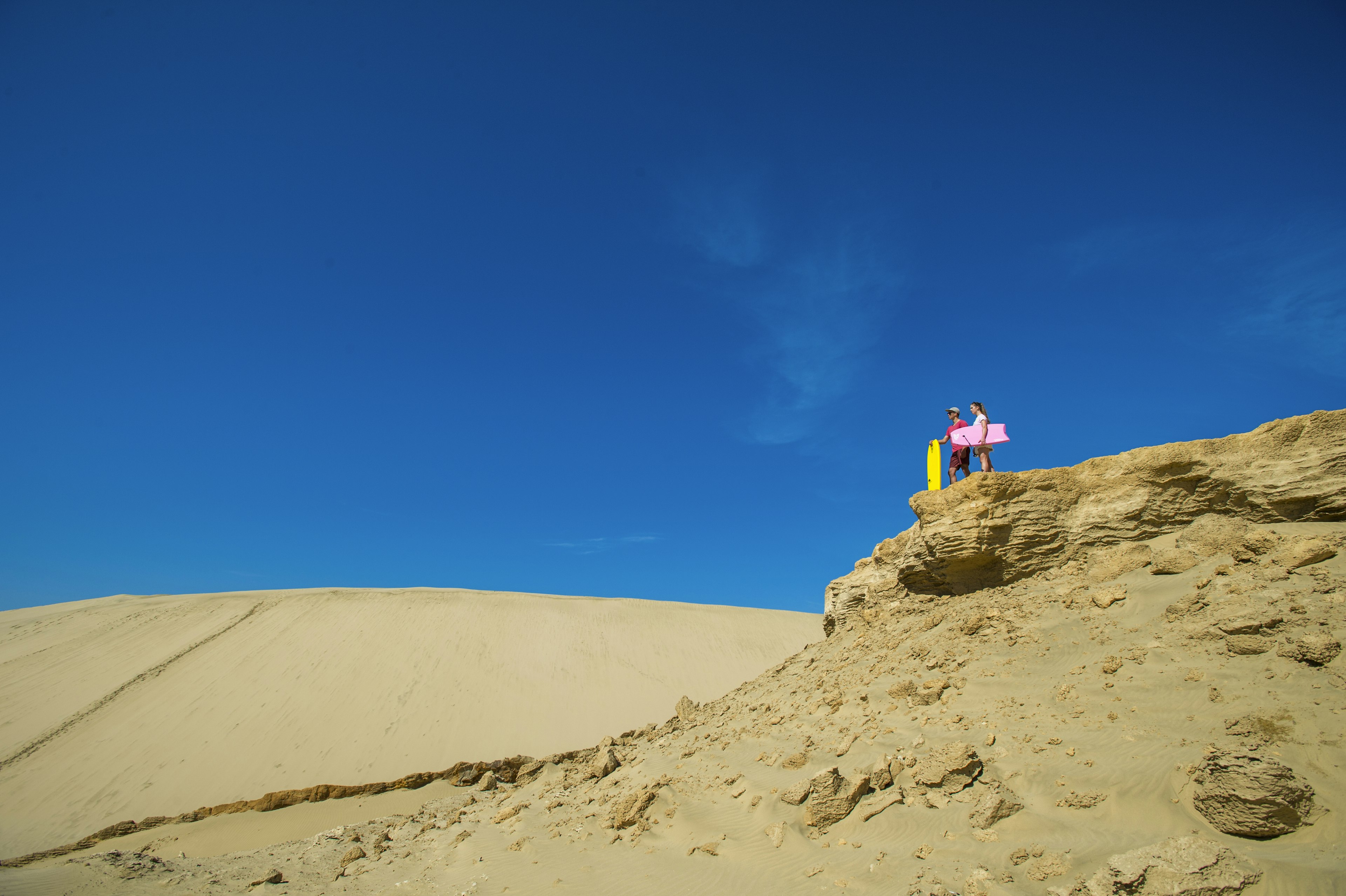 A pair of people stand at the top of dune, looking out on the arid landscape holding brightly colored sandboards.