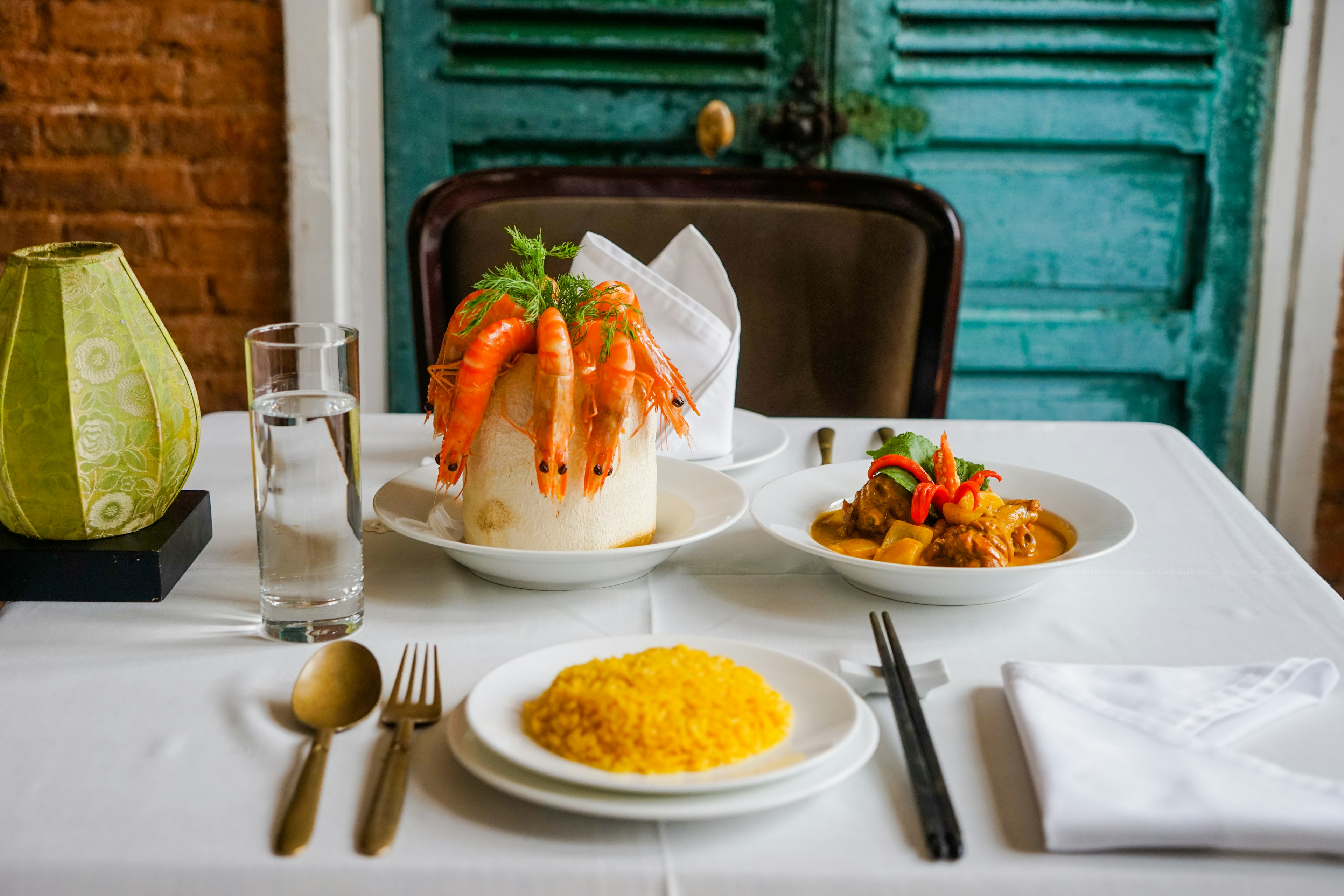 A table is set with dishes, cutlery and a glass of water. One dish is filled with yellow rice, another filled with vegetables and meat in an orange sauce and third has cooked shrimp dangling from a circular dish.