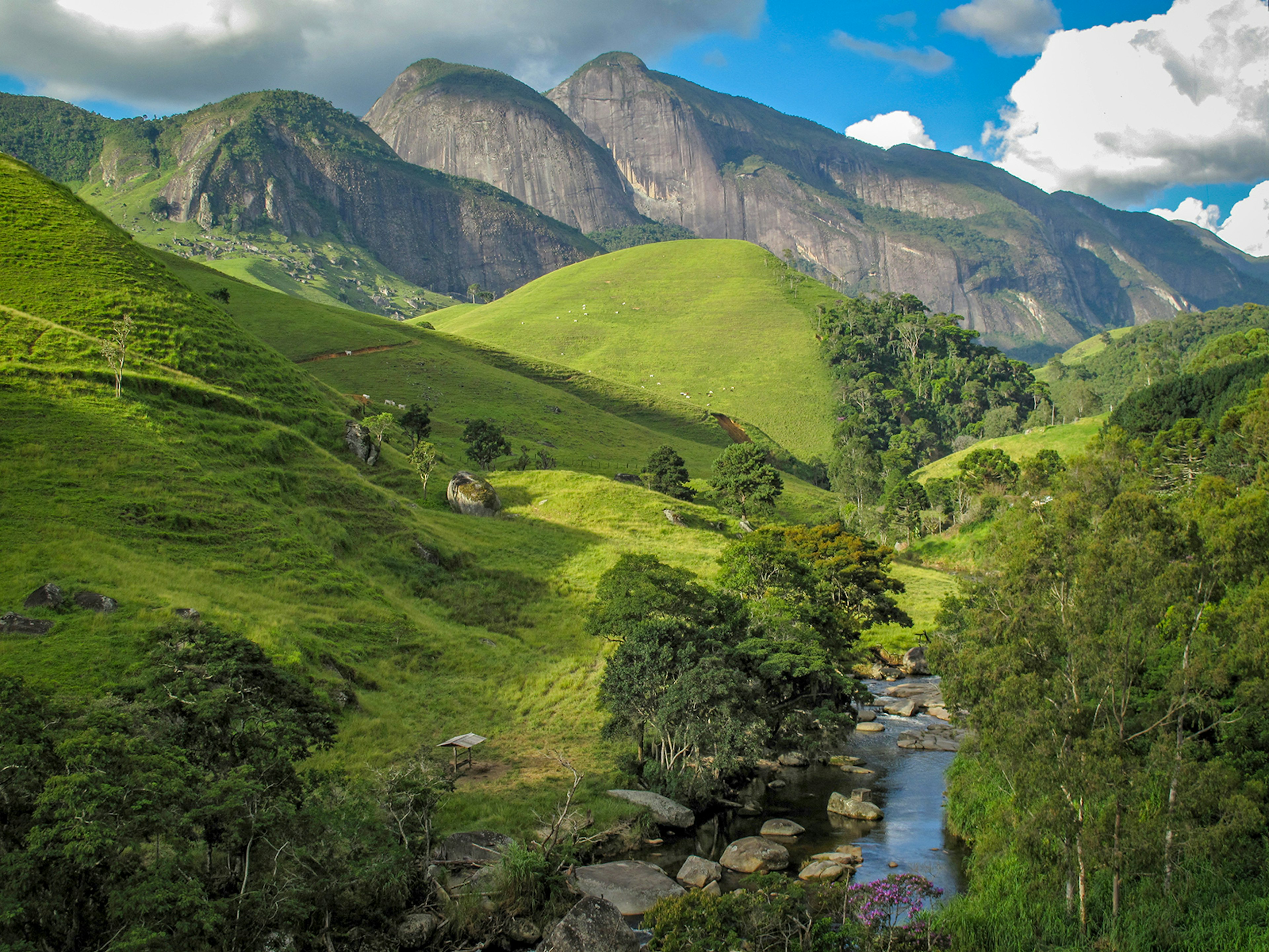 Rolling green hills in the foreground, framing a stream, with rocky mountains in the background © Phillippe Cohat / Getty Images