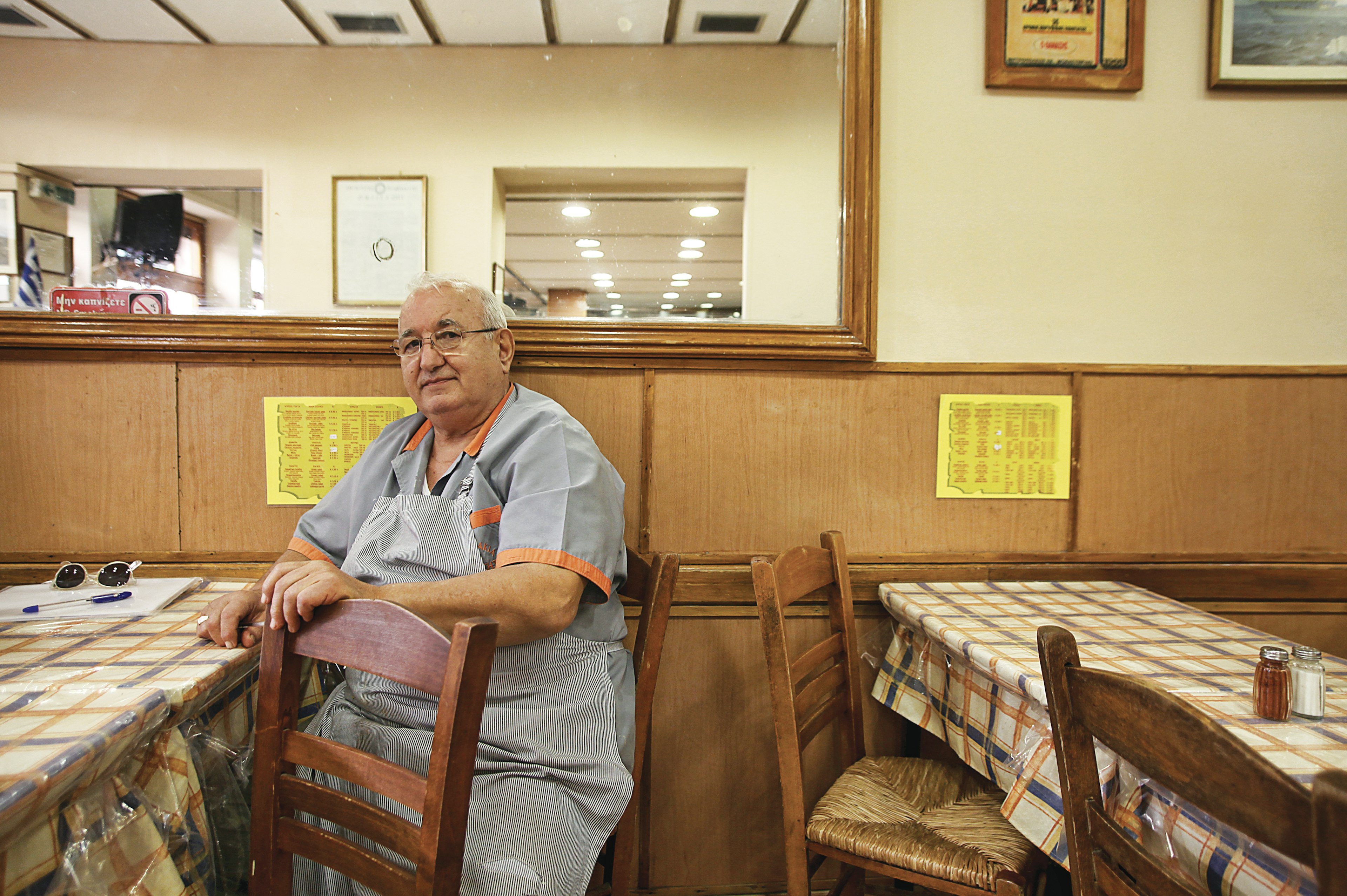 The owner of Thanasis restaurant sits at one of the tables, whcih are covered in blue and orange plaid clothes covered in clear plastic. He is wearing a light blue chefs shirt with orange trim on the sleeves and collar. The walls are paneled in wood and veneer, belaying the age of the restaurant