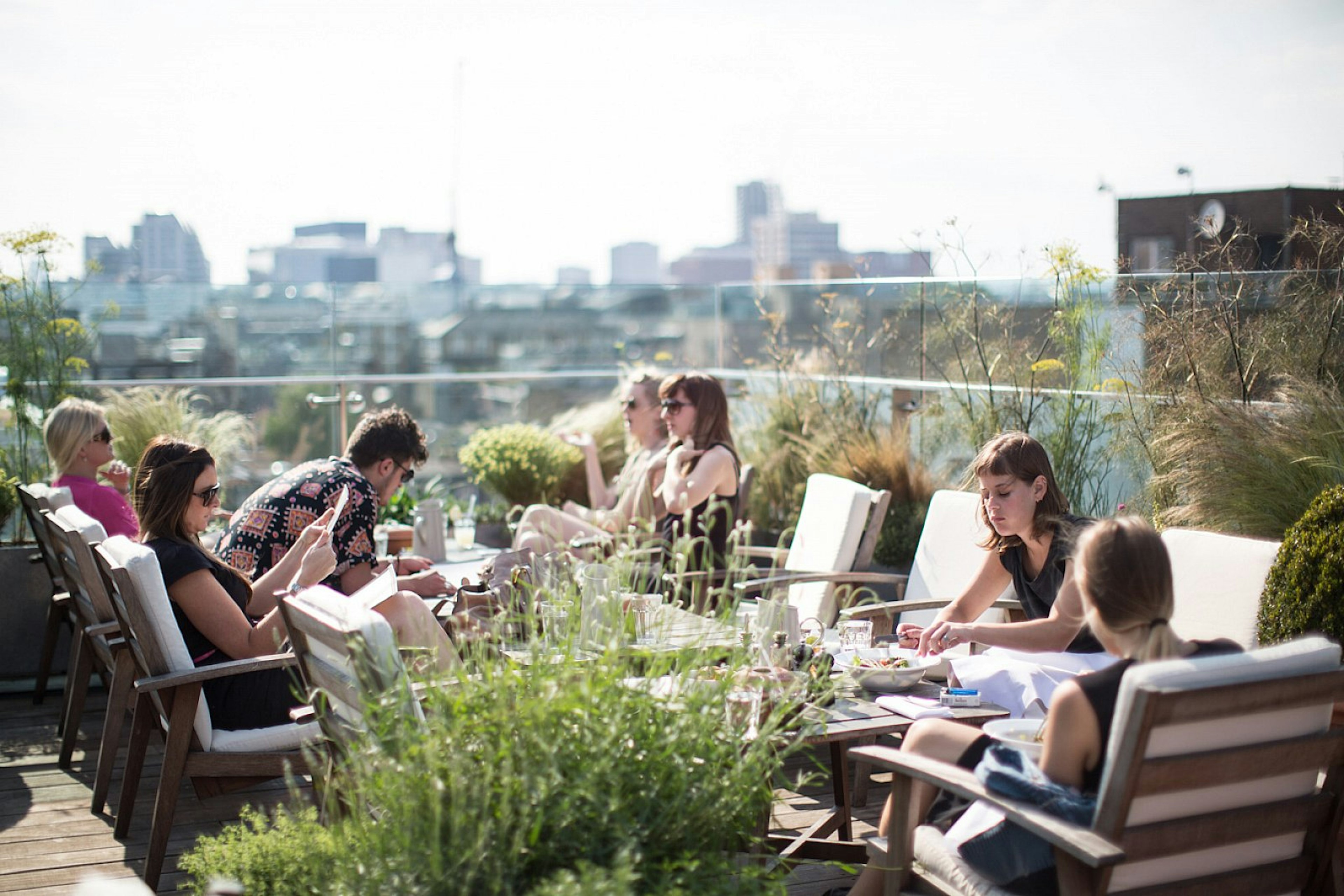 People sit at white chairs on the Boundary rooftop on a sunny day, surrounded by plants; a blurred city skyline is visible beyond.