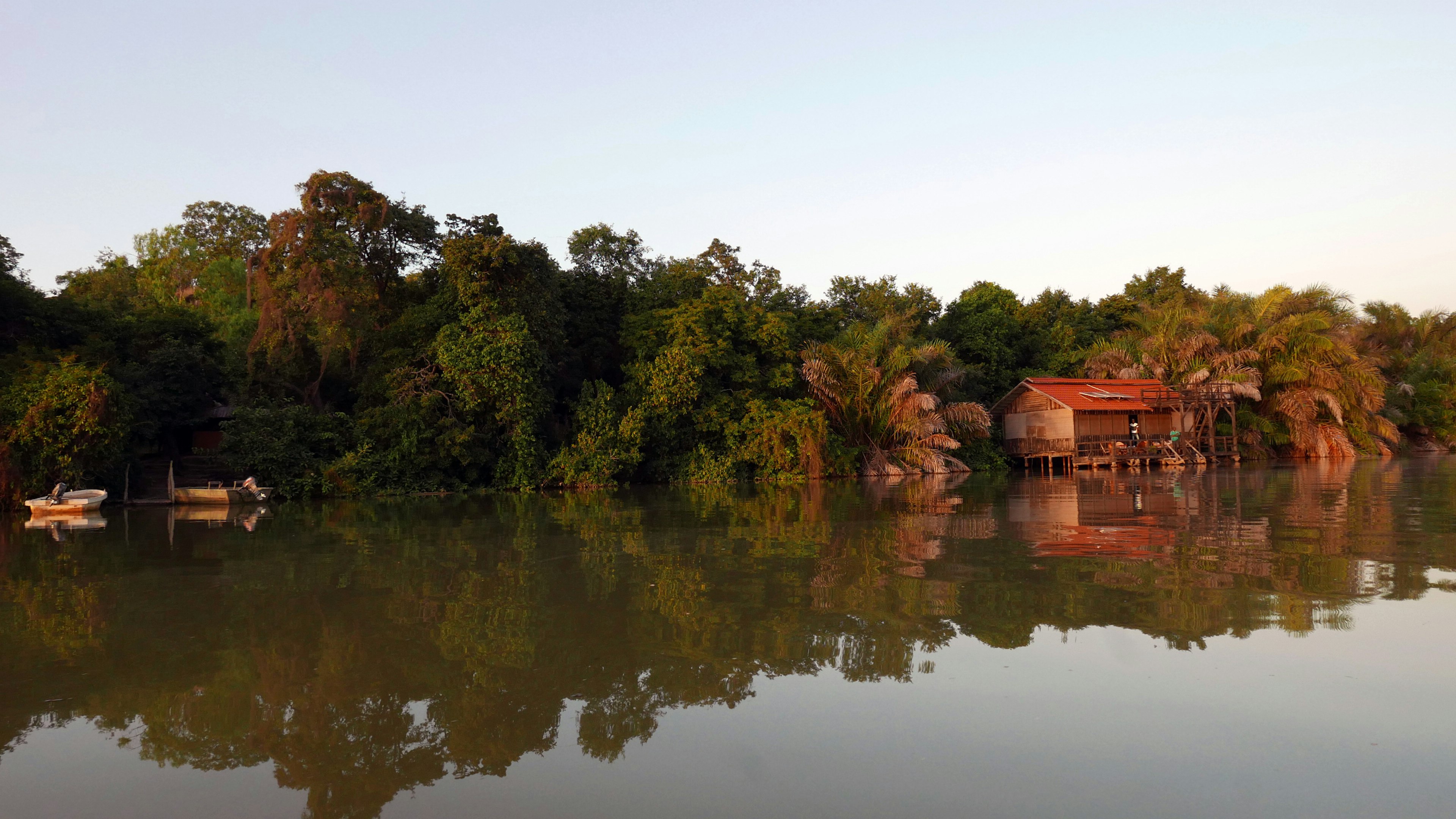 A forested section of riverbank reflects in the mirror-like surface of the River Gambia; nestled into the trees is a red-tinned roof wooden building - nearby are a couple of small boats moored on shore.