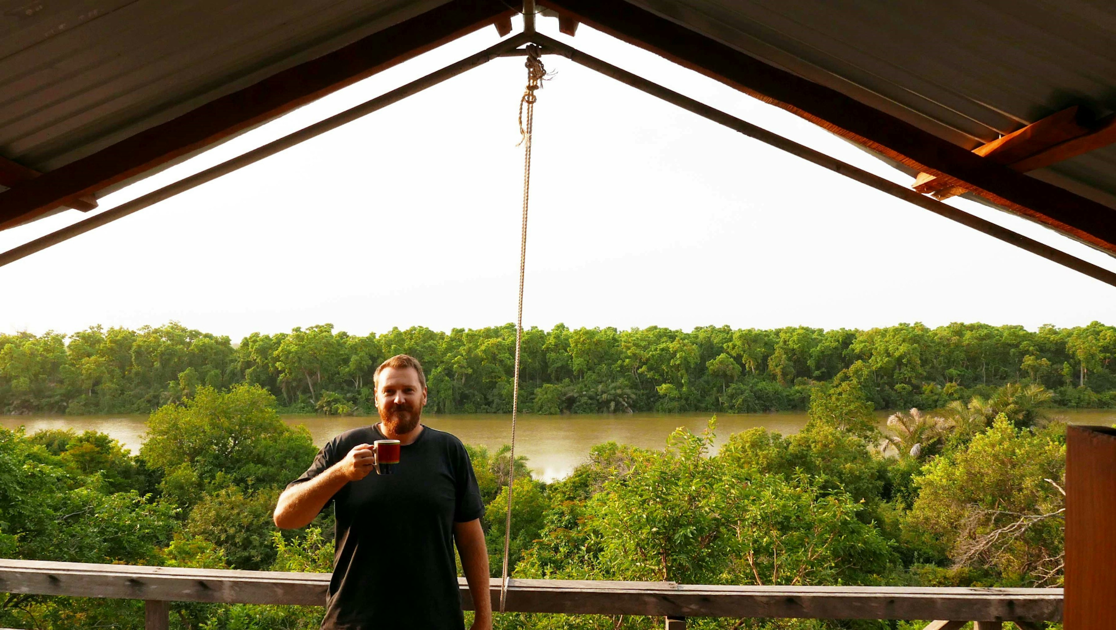 Bearded Joseph holds up a cup on the edge of his tent; behind him forest lines both sides of the River Gambia.