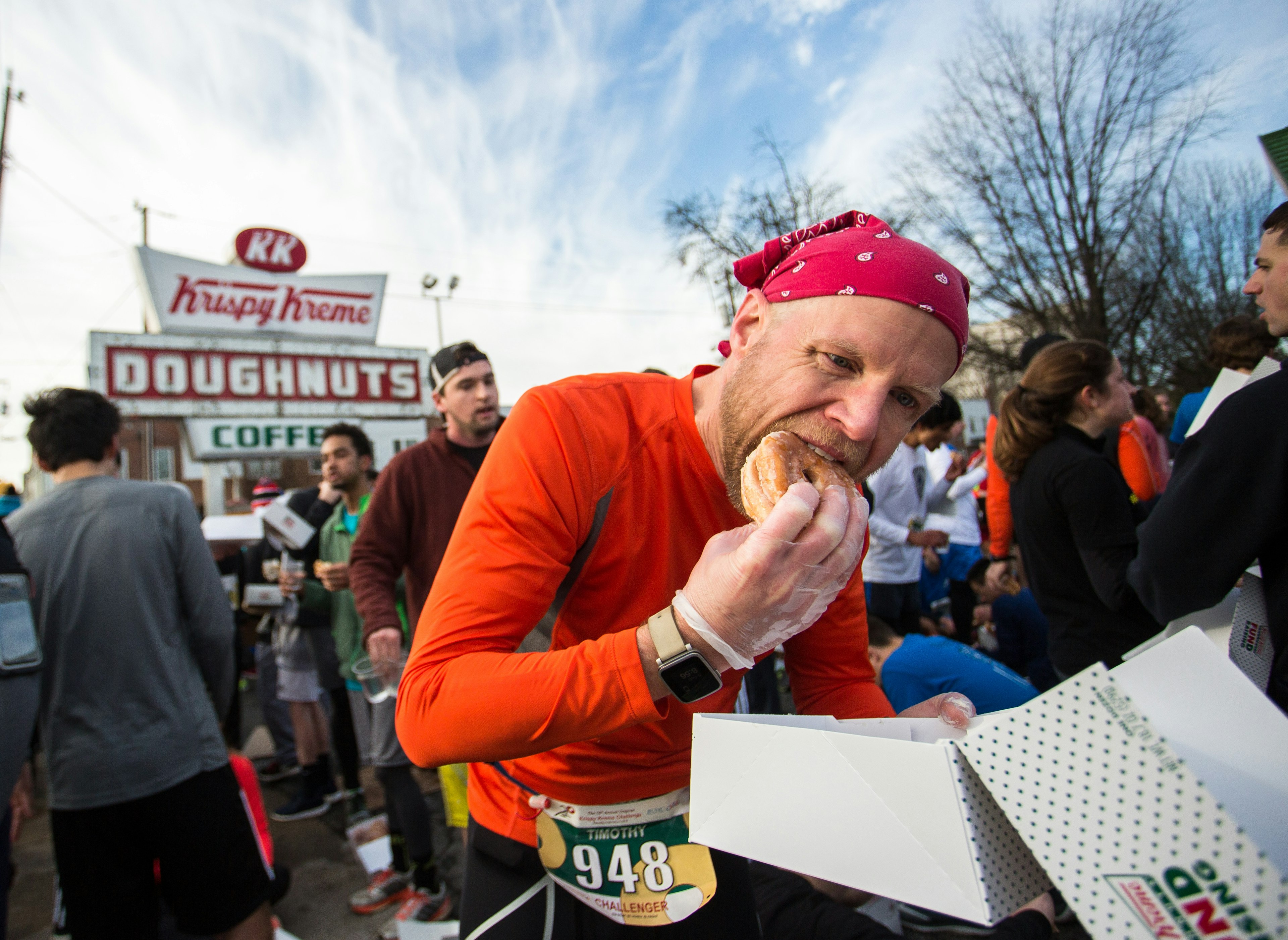 A man shoves a doughnut into his mouth during a run. In the background is a Krispy Kreme stand