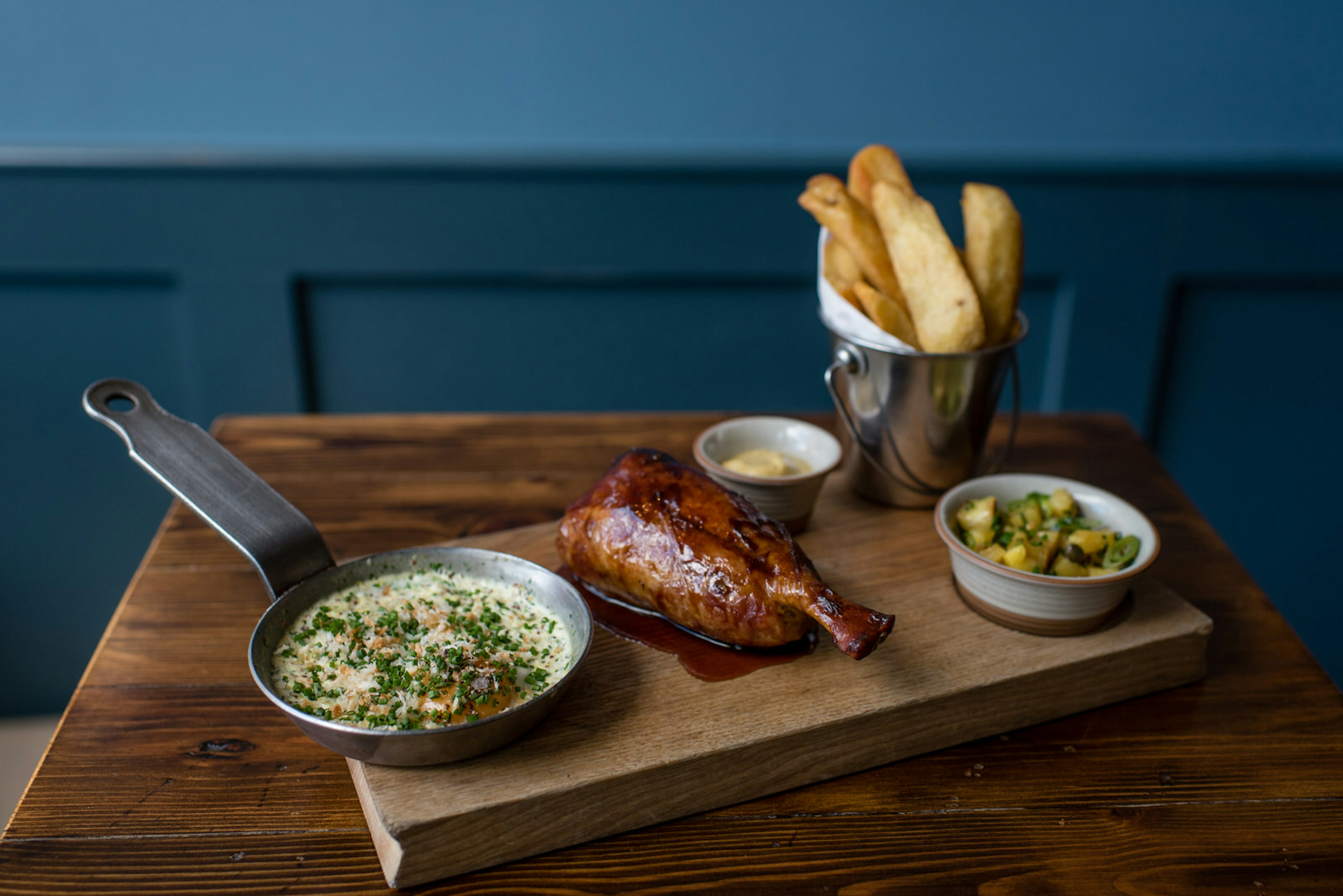 A close-up of ham, egg and chips served on a wooden cutting board. The egg is still in a miniature steel frying pan and the chips are in a small tin bucket.