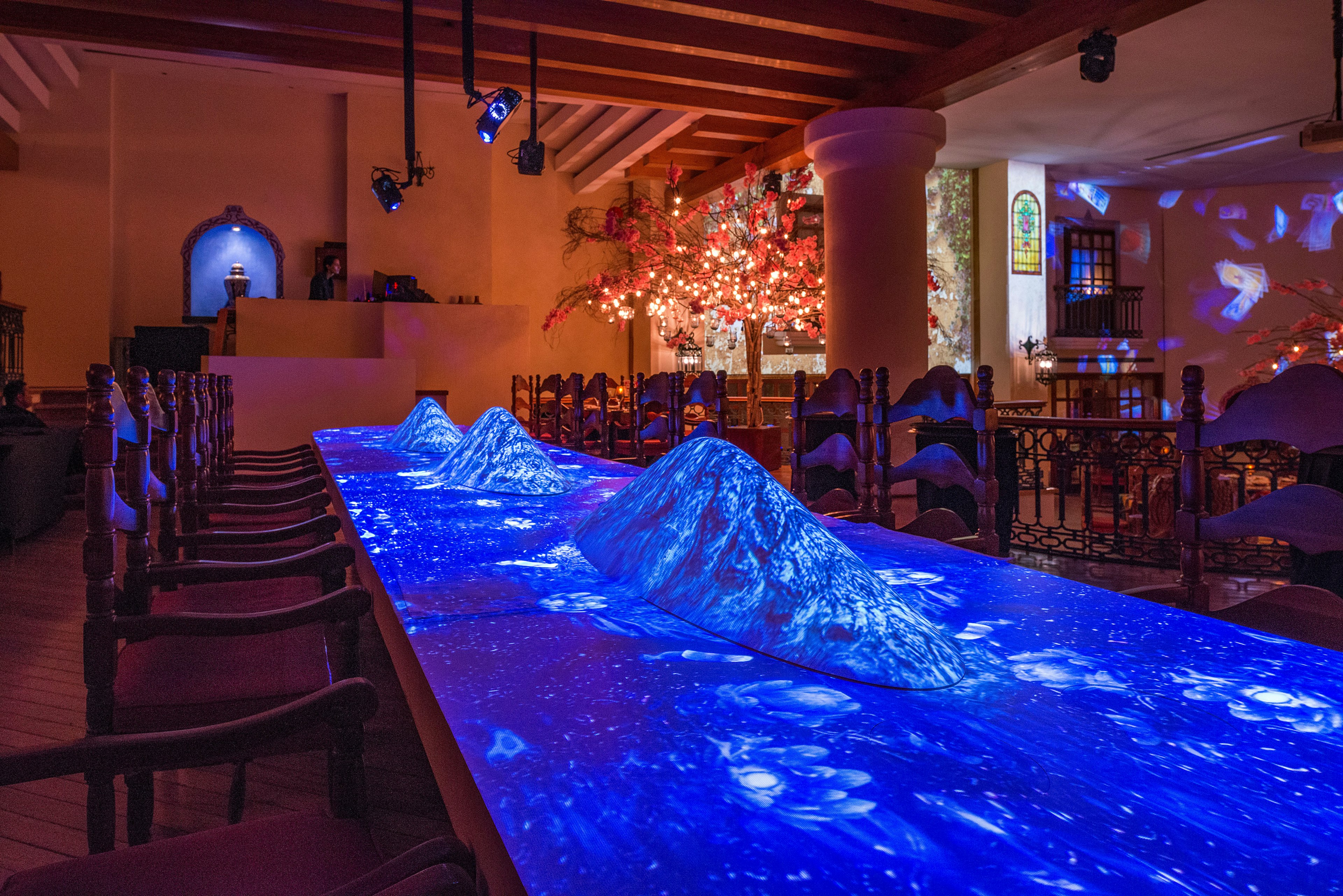 Three raised structures spaced out on a large table are covered with a blue and white light from an overhead projection.