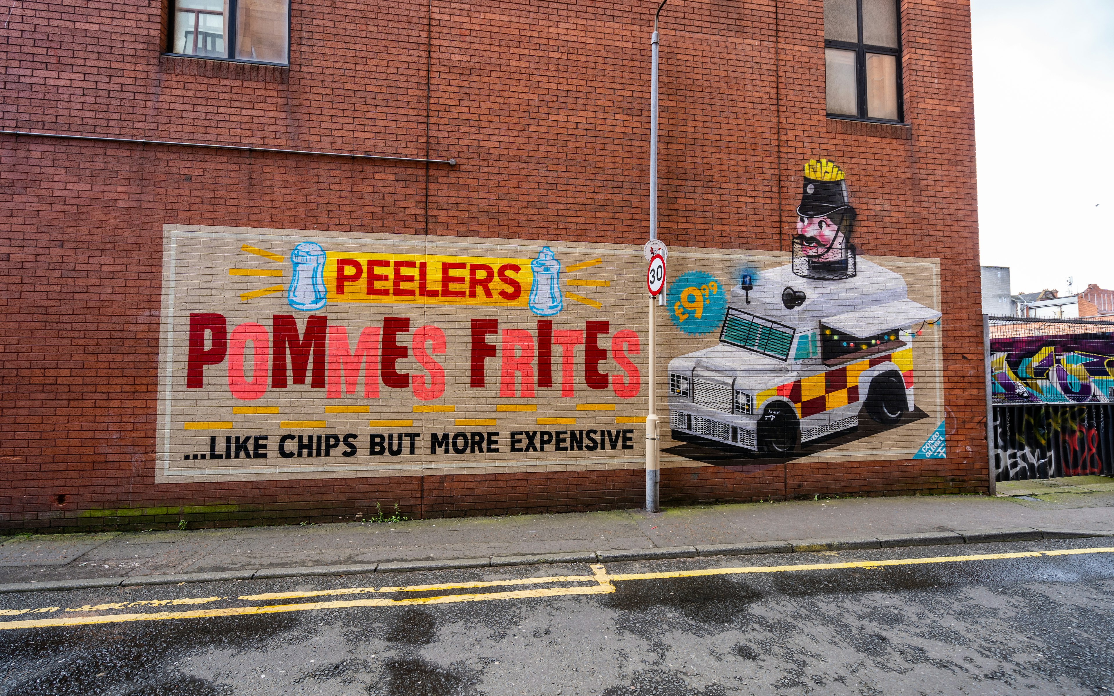 A red brick wall, painted with a colourful mural depicting an advertisement for Peeler's Pommes Frites in the style of a vintage poster.