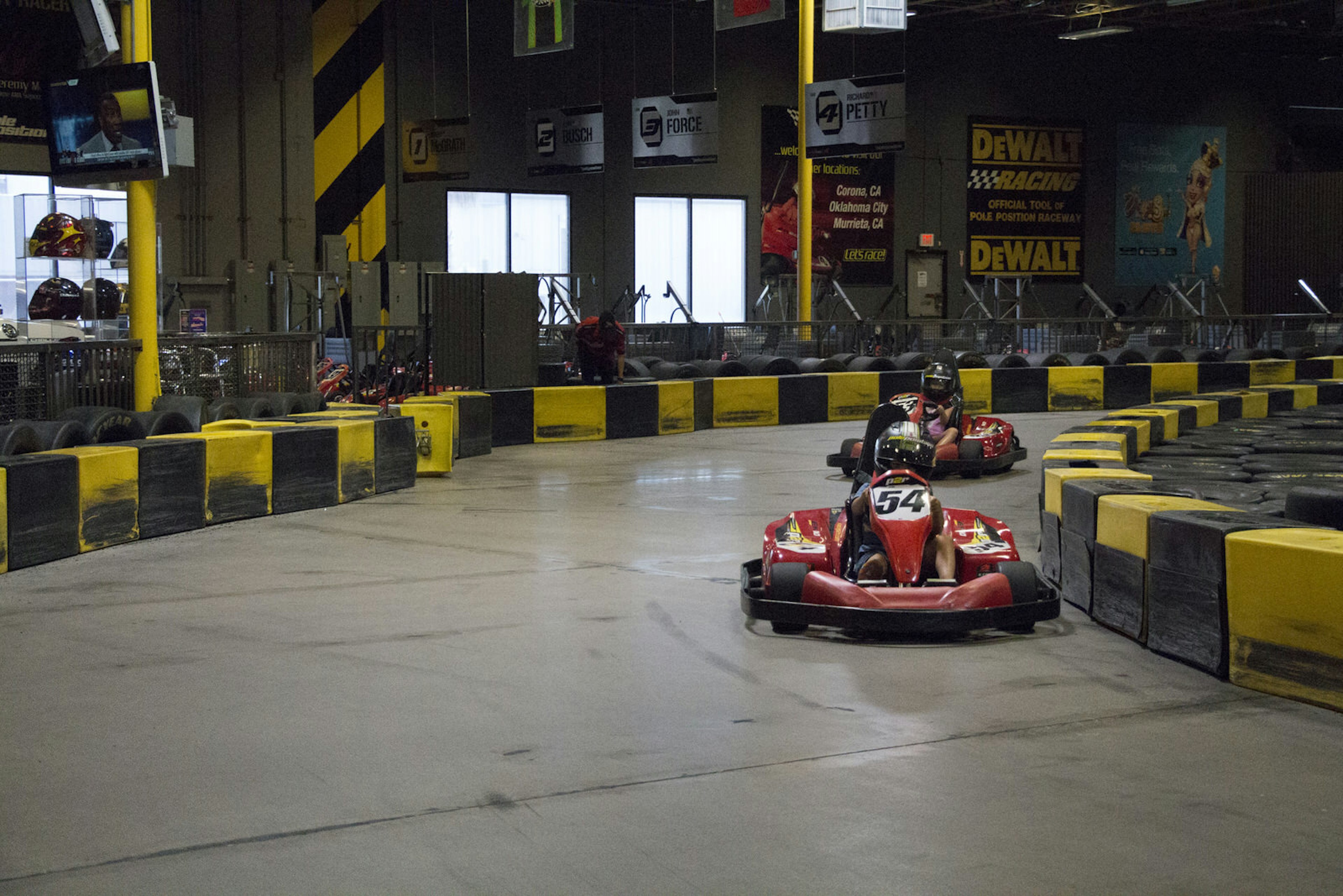 Yellow and black barriers border the wide expanse of a twisting go-kart track, where riders in red cars speed around a corner.