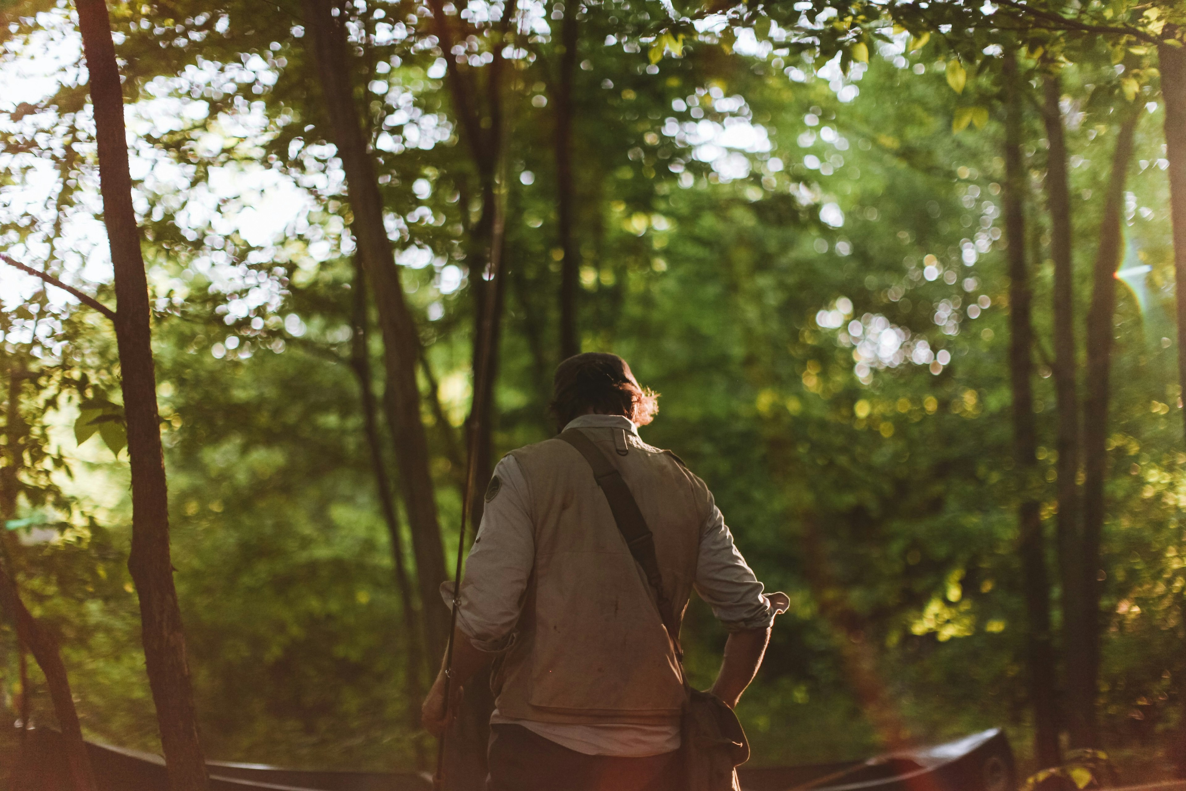 Co-founder Tom walks through the woods near Livingston Manor Fly Fishing Club, with his back to camera. He holds a rod in his hand.