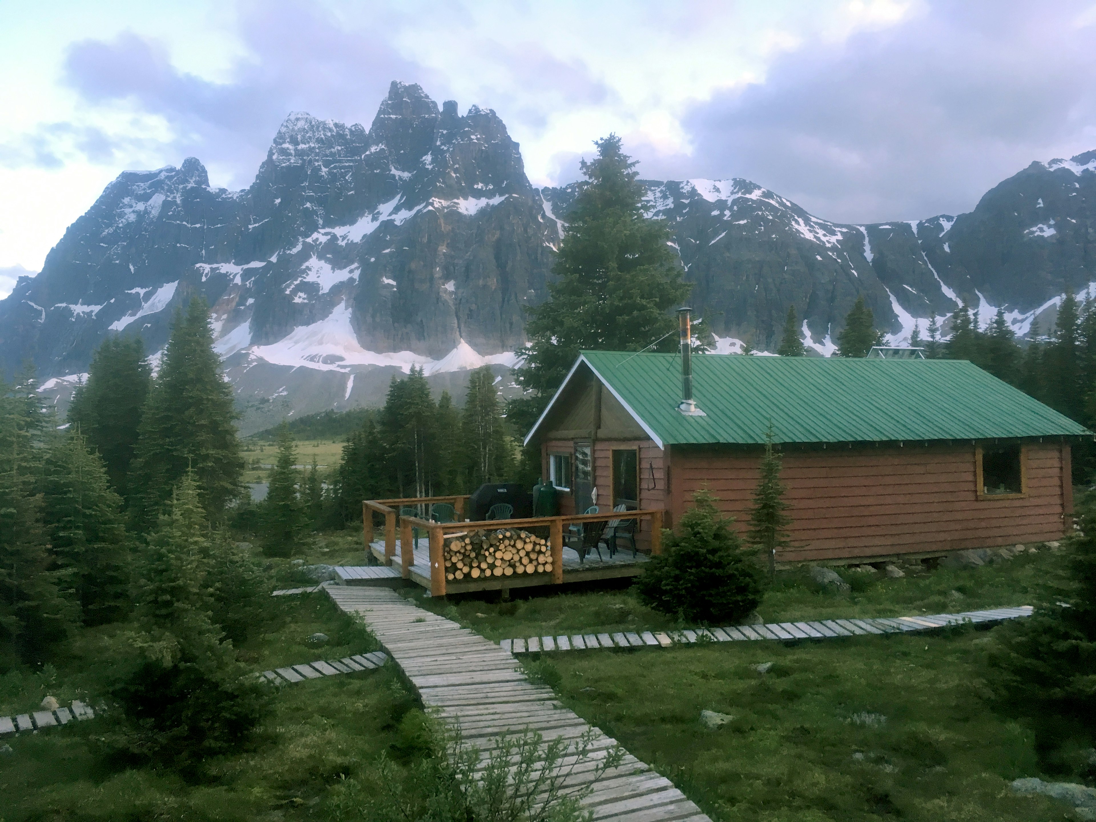 A green-roofed lodge with a large deck is seen in the shadow of a large snow-covered mountain; Banff and Jasper's backcountry lodges