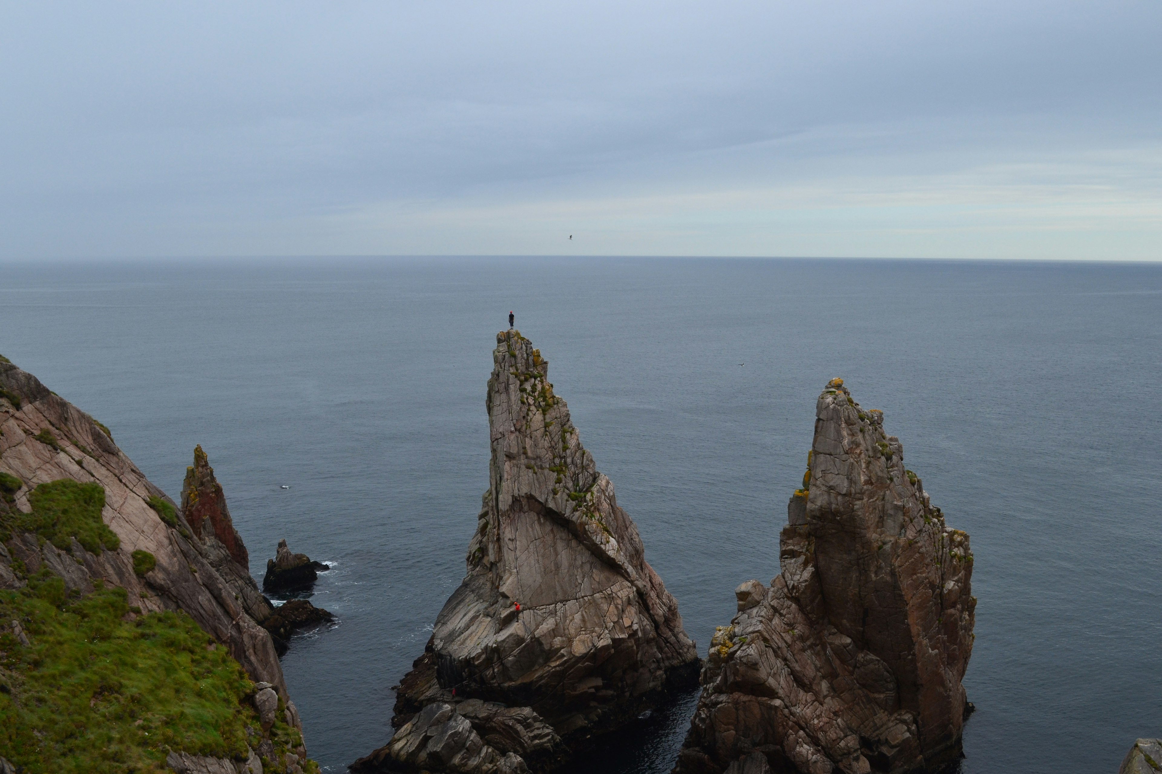 A person stands on the top of high, narrow piece of rock jutting directly up from the sea.