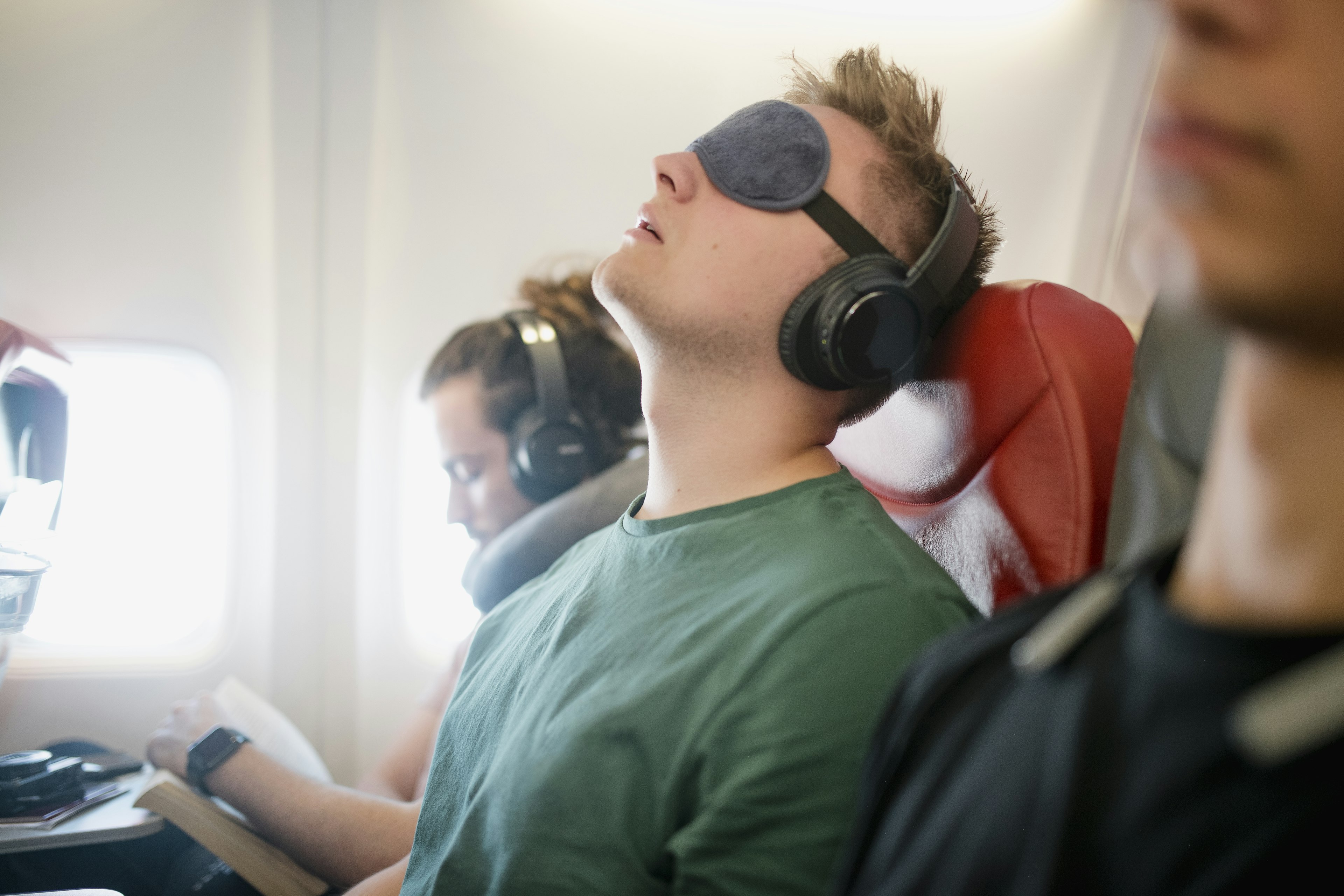 A man leans back in an airplane seat wearing an eye cover and headphones. Next to him is another man with headphones and neck pillow.