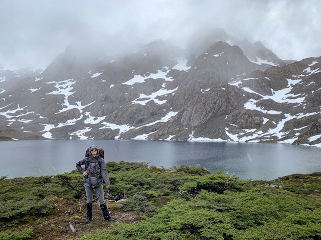 A hiker poses in front of a lake backed by snow-lined mountain slopes; sleet or rain falls