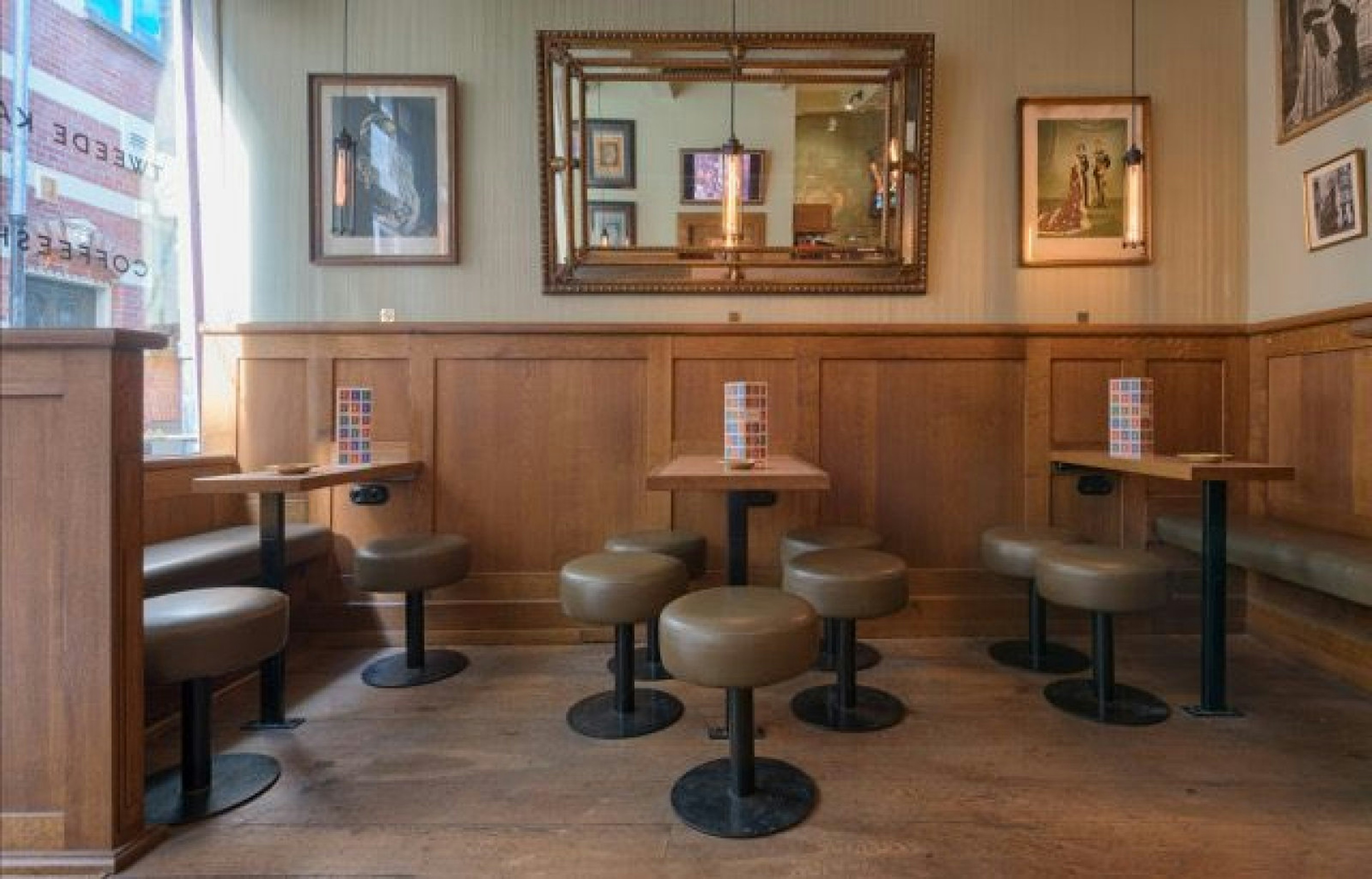 The interior of Tweede Kamer coffeeshop, Amsterdam. The walls are hung with a large ornate mirror as well as images of royals. There are three wooden tables with stools and couches around them.