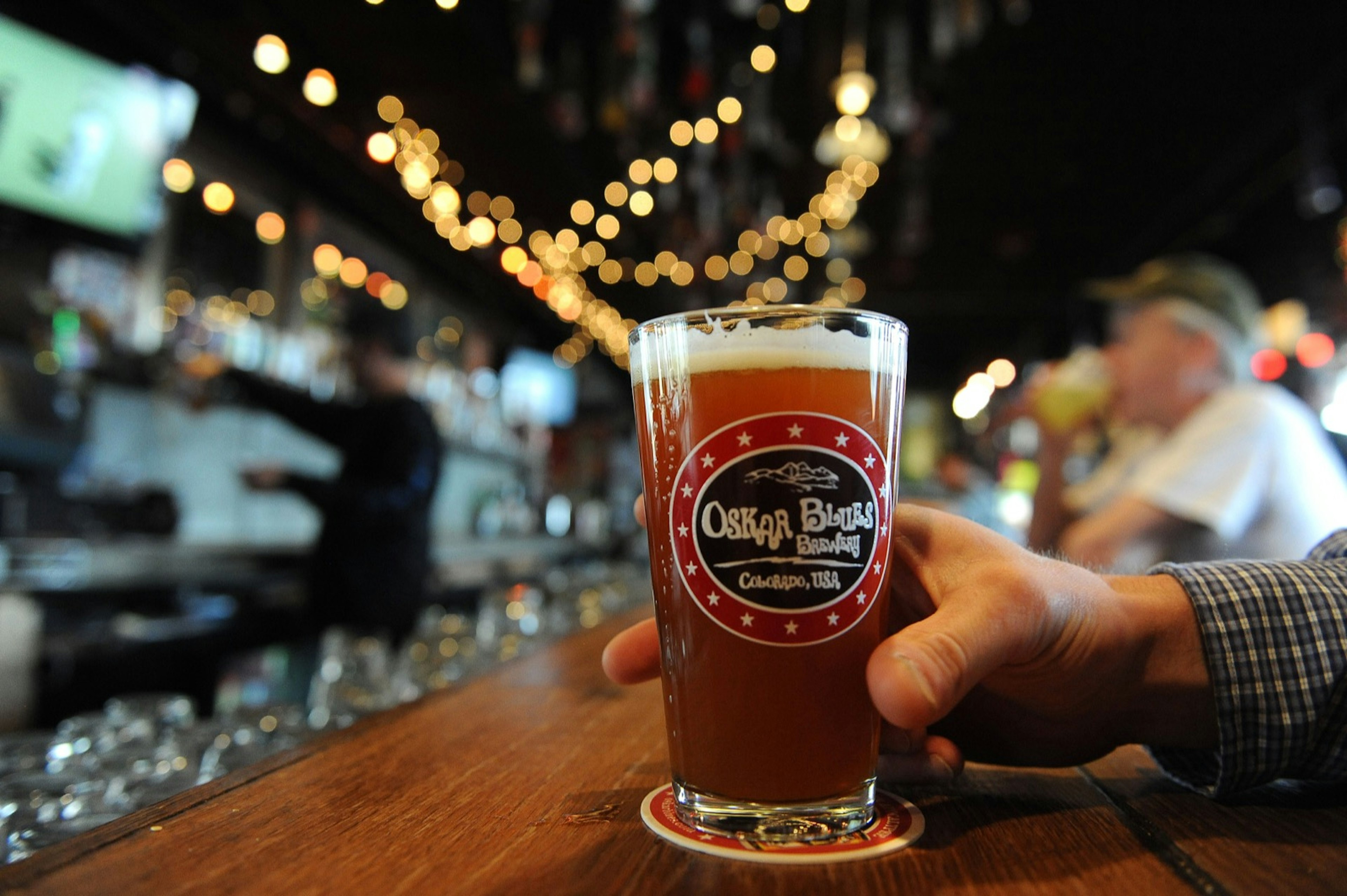 A man holds a glass of amber-colored beer resting on a circular coaster on a wooden bar. A collection of patrons drinking beer can be seen in the background. Oskar Blues is among one of the top craft breweries in the country.