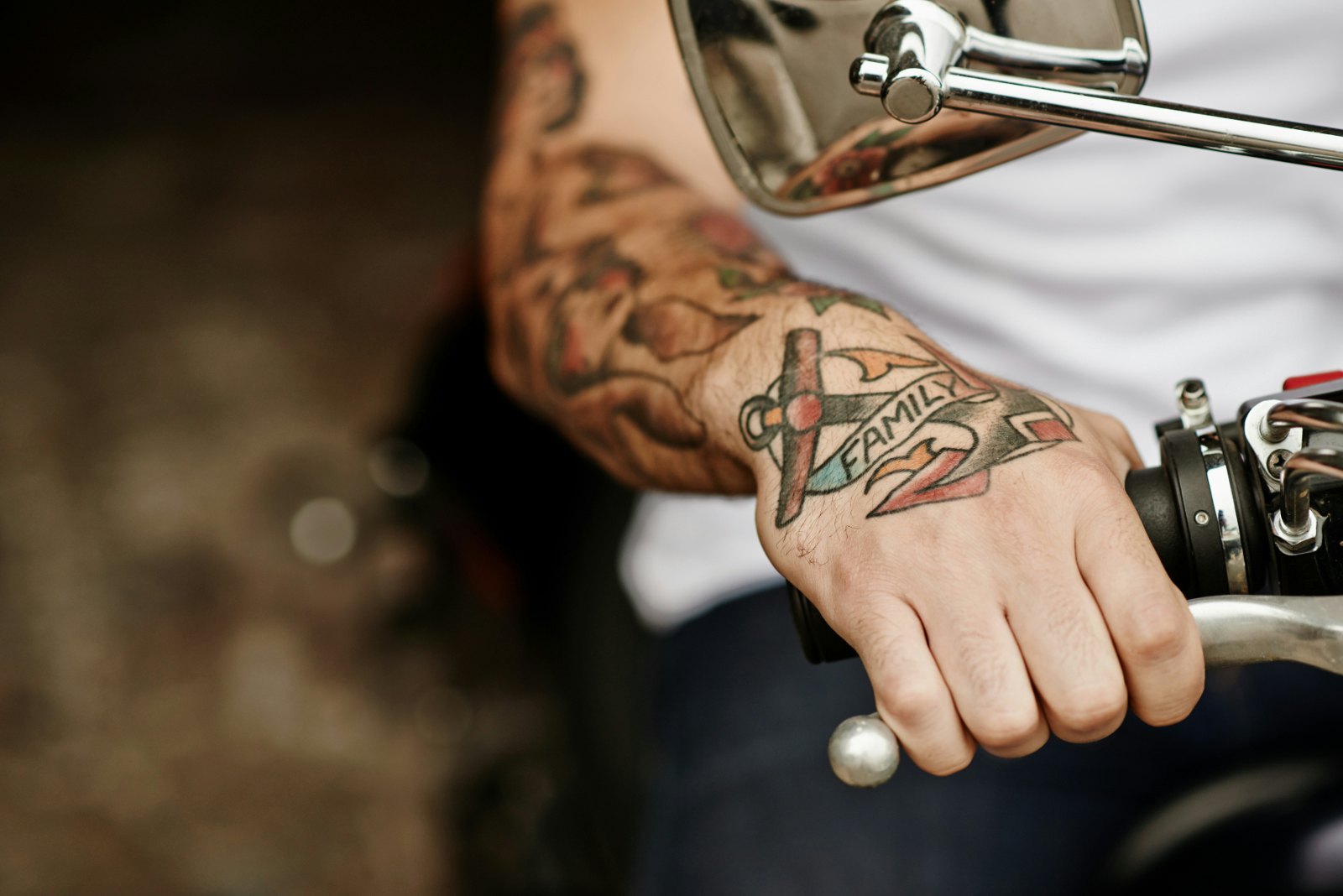 A close up of a person's tattooed hand on the handle of a motorbike. The tattoo on their hand says 'Family' and they have a sleeve tattoo up their arm.