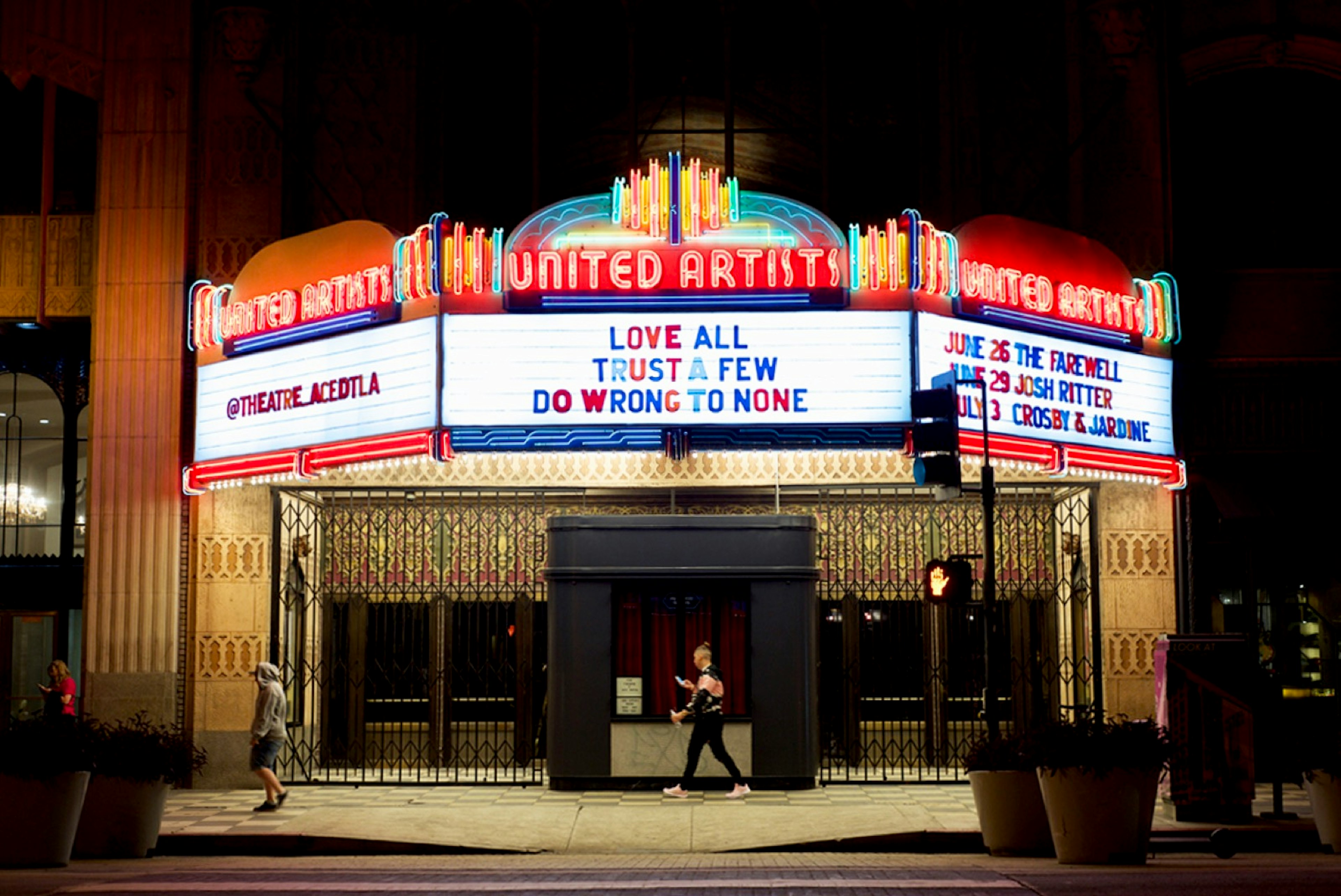 People walk in front of the Ace Hotel at night, which has an old-fashioned movie house marquee lit up in neon; Los Angeles neon