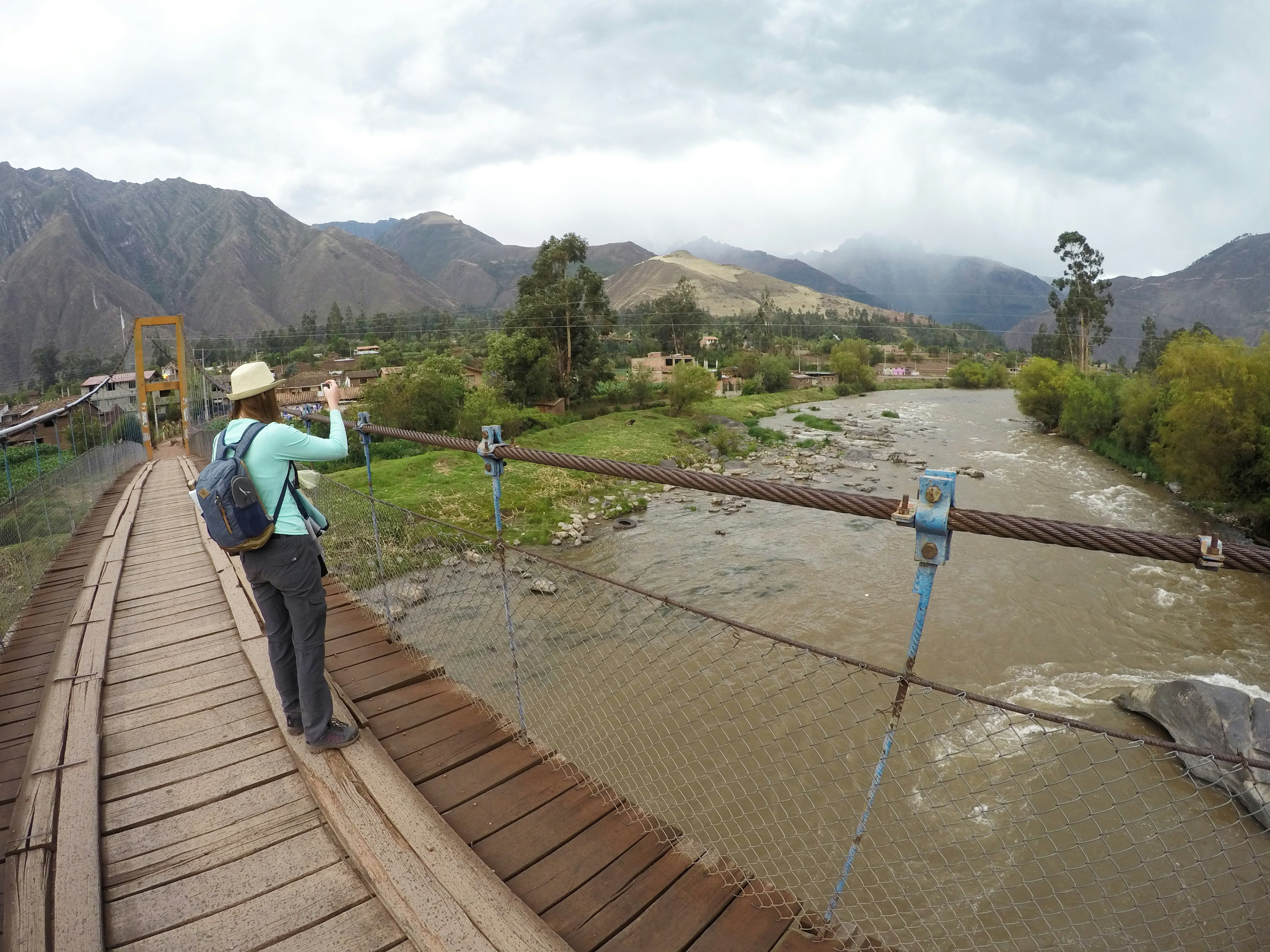 A woman stands on a bridge taking a picture with her camera, overlooking a river with a mountain covered in mist in the distance