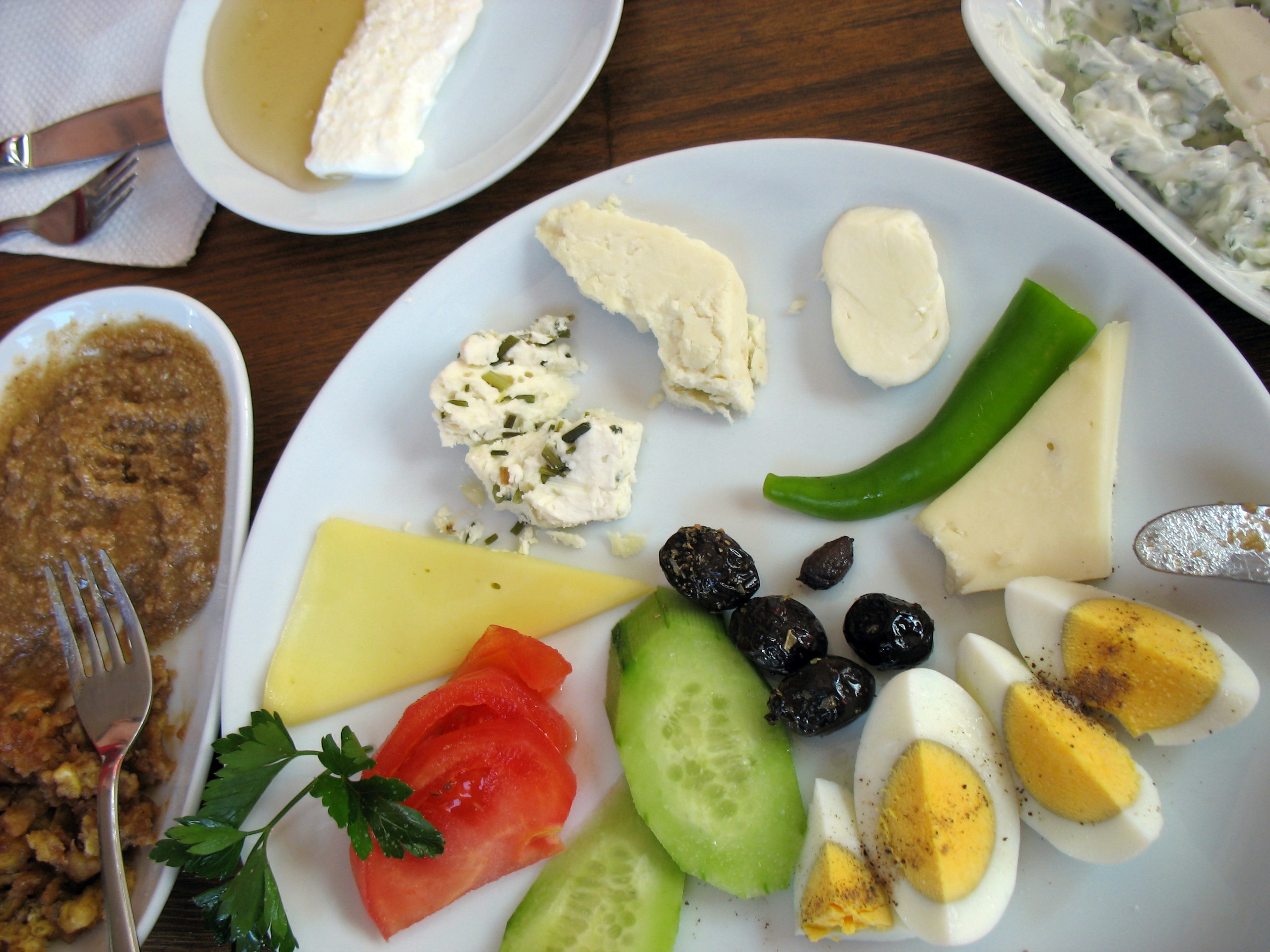 A plate of food is filled with a sliced boiled egg, slices of cucumbers, olives, cheese and tomatoes. There are plates filled with different types of sauces around the main plate.