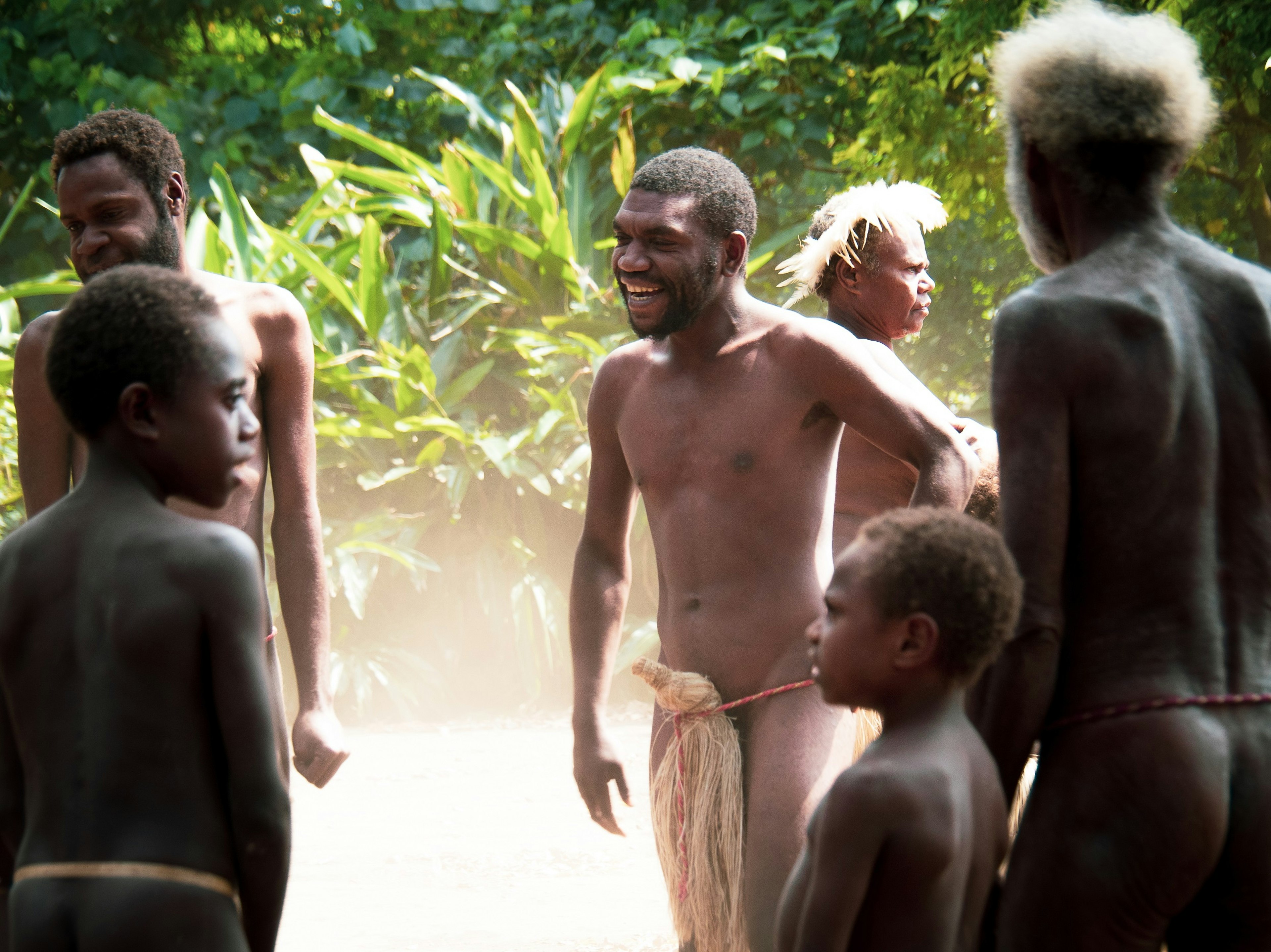 A group of men from the village of Yakel stand facing each other in a circle of sorts; they are just wearing a string around their wastes, with a narrow bunch of dried grass over their groins; behind them is lush vegetation and there is a lot of dust around ground level, which is likely from dancing.