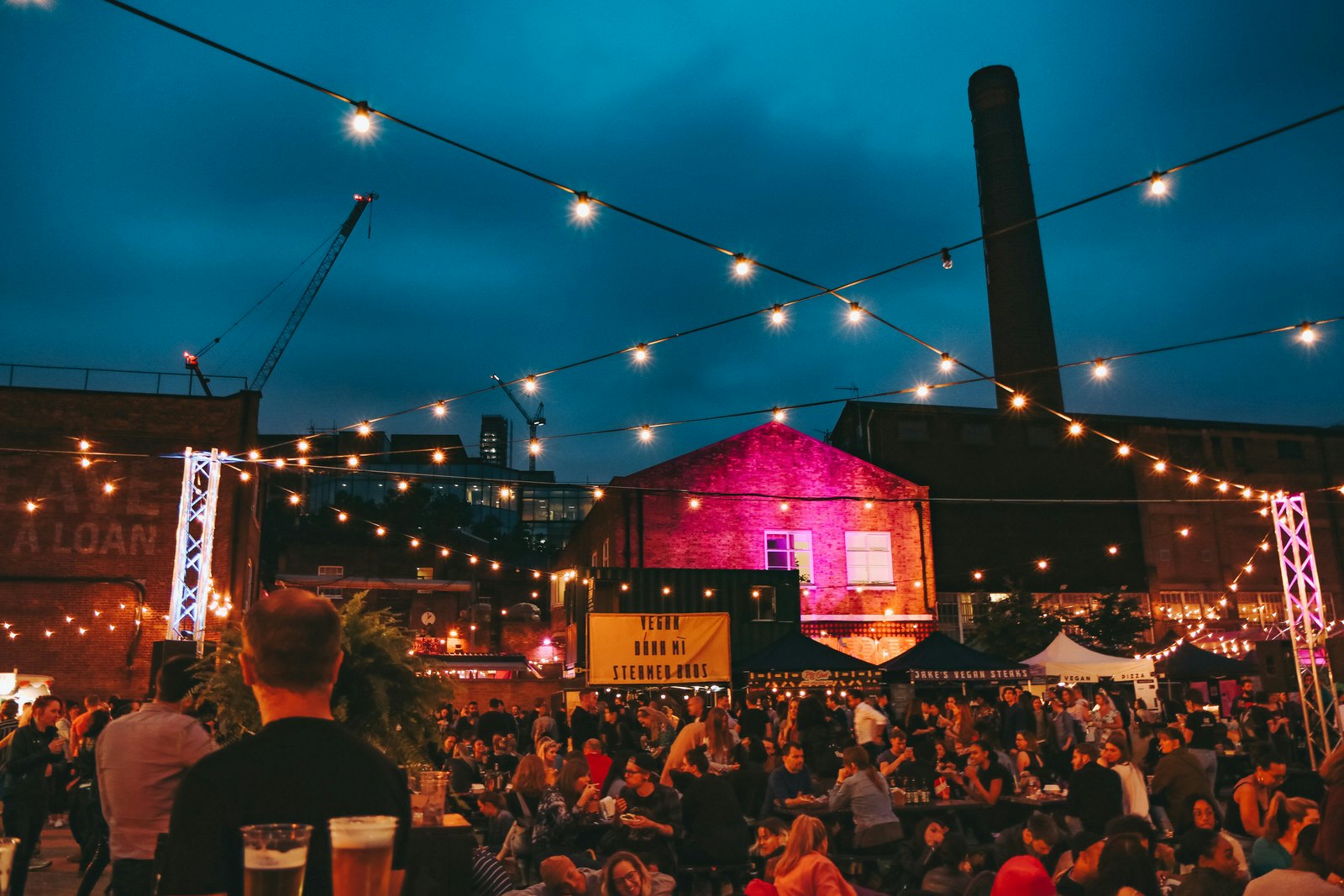 A crowd of people are eating and drinking at night-time in a square near Brick Lane; there are vegan food stalls along one side of the square and a factory chimney and modern office buildings in the background.