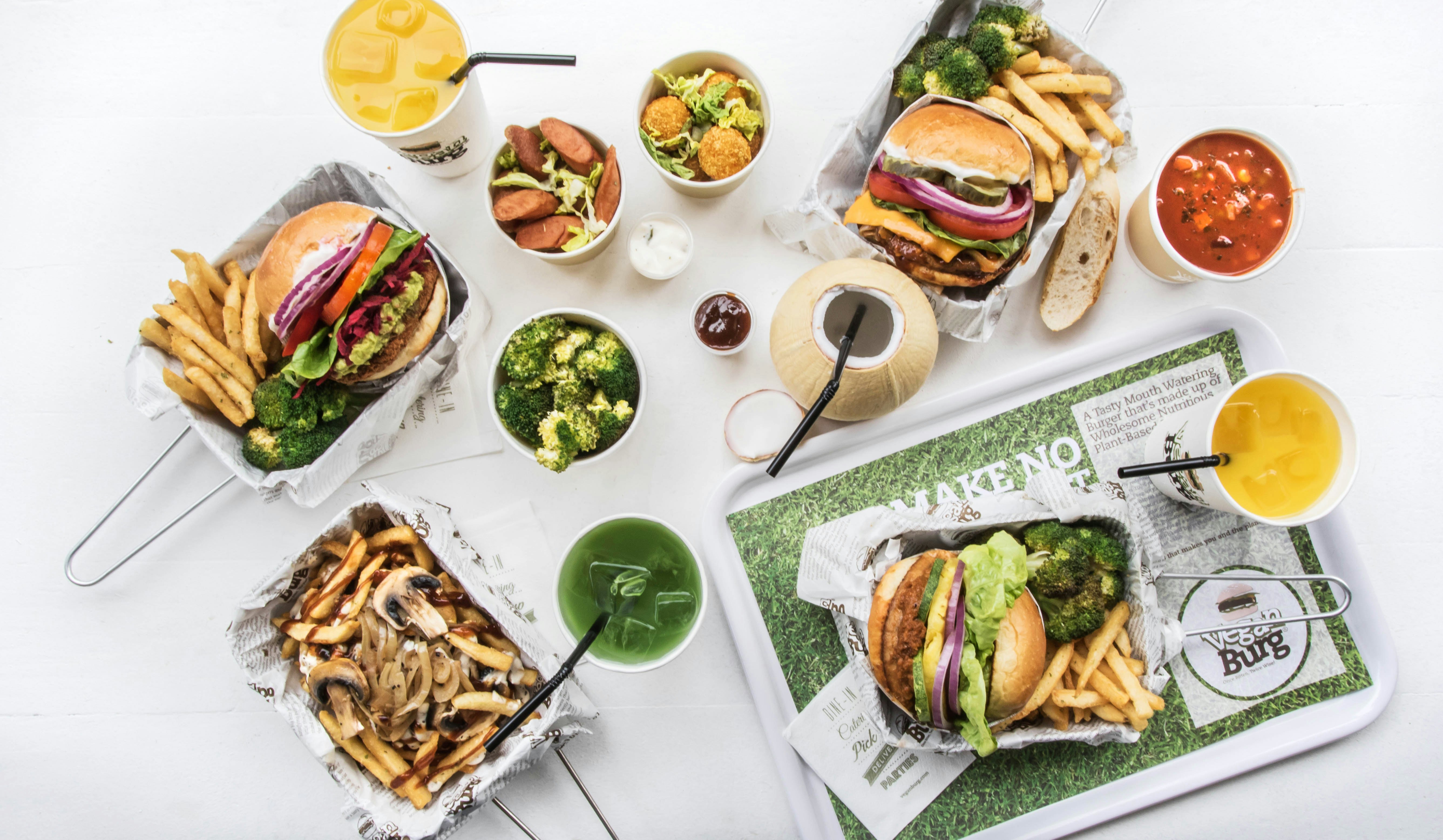 An overhead view of a table filled with dishes of vegan burgers with broccoli and french fries, small salads and a variety of cups; San Francisco Bay vegan restaurant