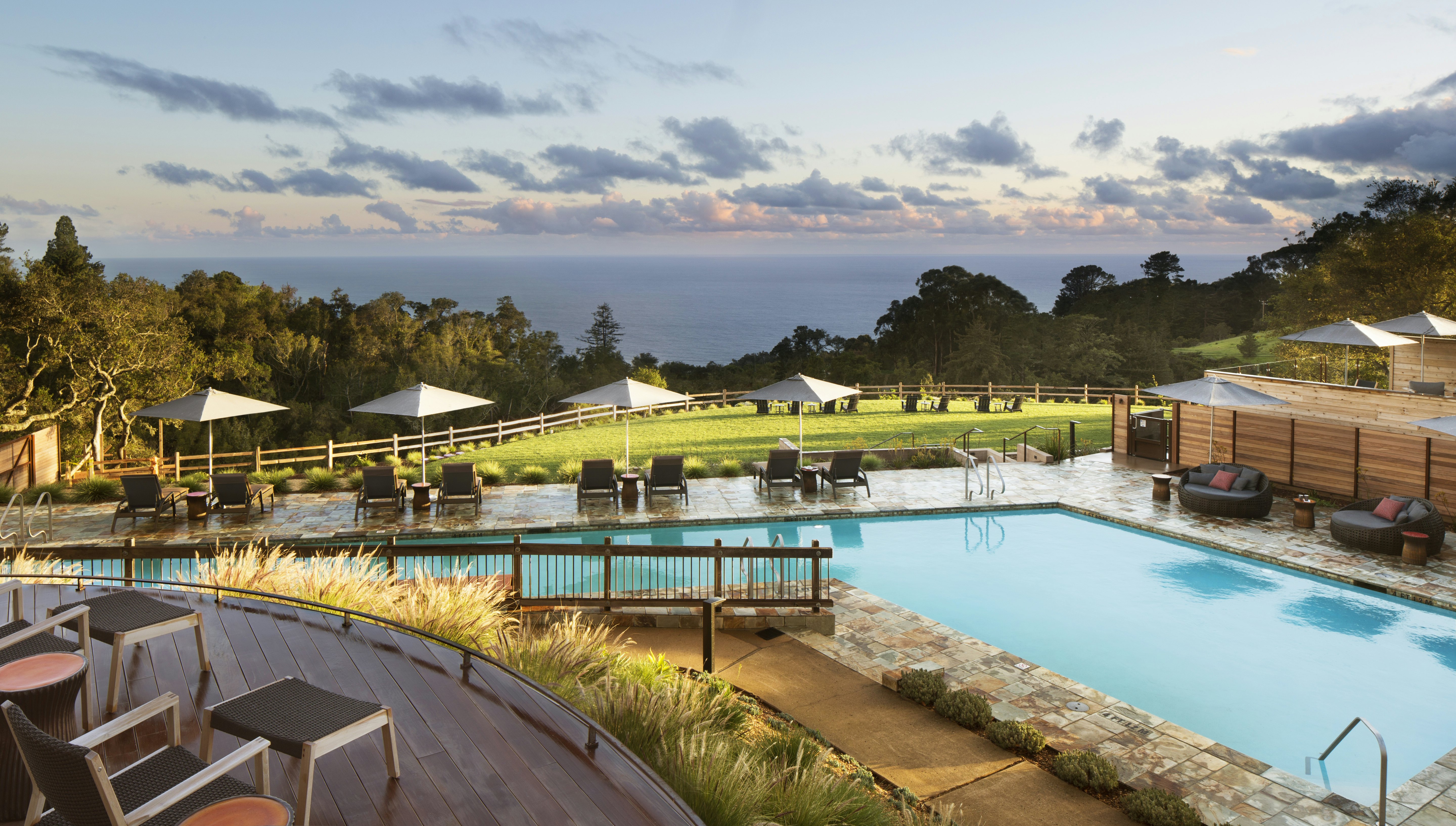 A quirkily shaped swimming pool with stylish pool furniture arranged around it, with trees and the Pacific Ocean beyond.