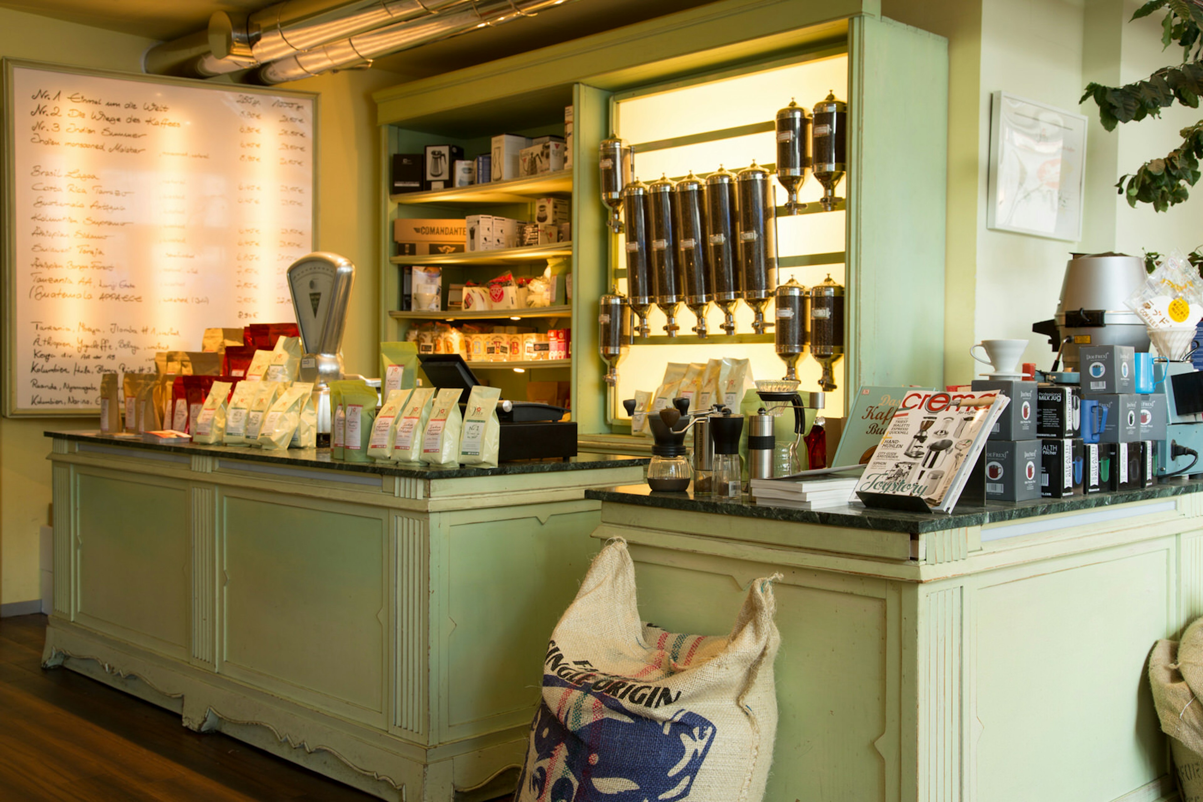 The interior of Vits der Kaffee, roastery and cafe. The walls and marble-topped counter are painted a very pale green. The counter and shelves behind it are stacked with coffee products and there is a whiteboard to the left displaying the different blends of coffee available.