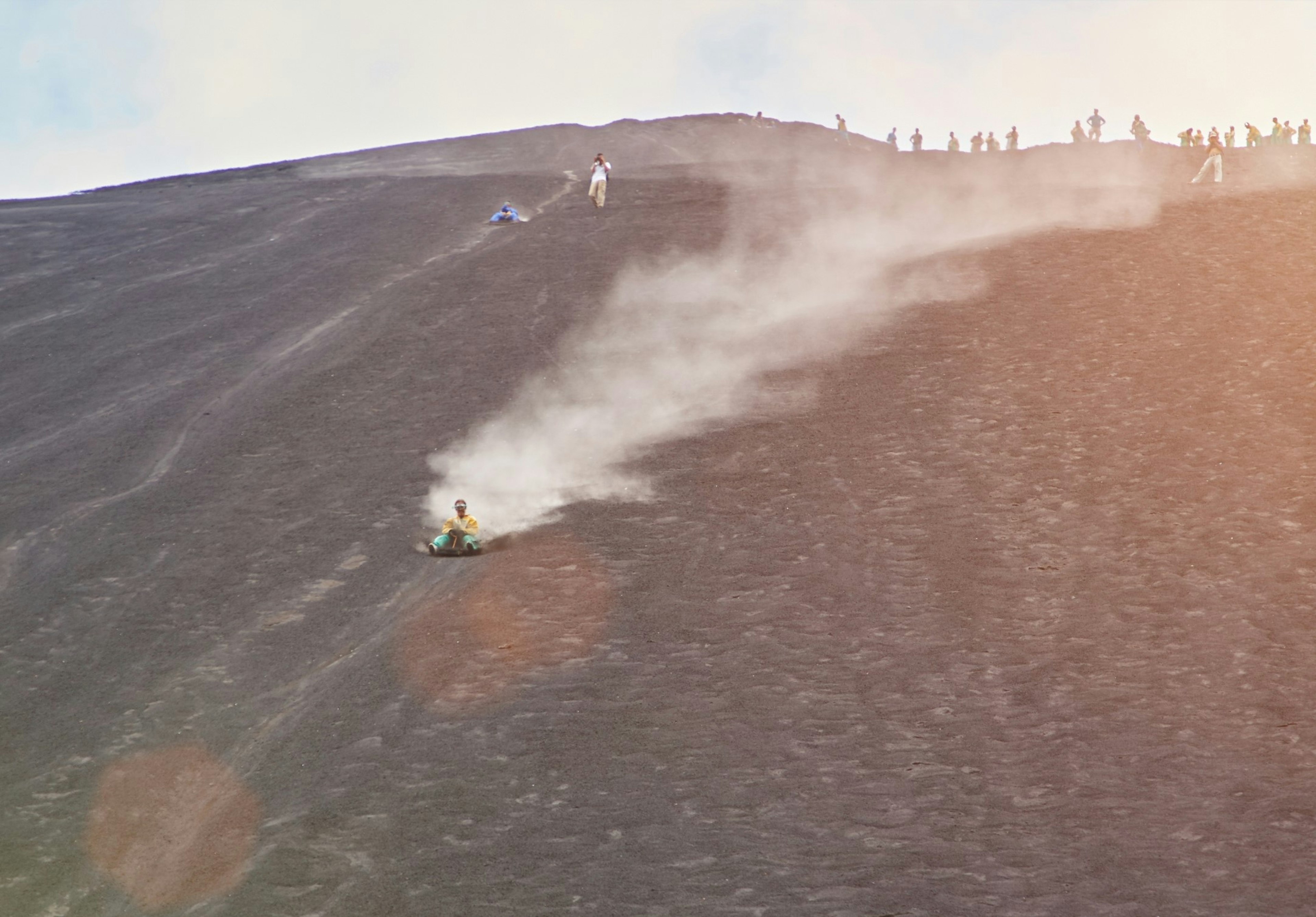 A man is sliding the side of a steep hill, on a volcano, while sat on a board.