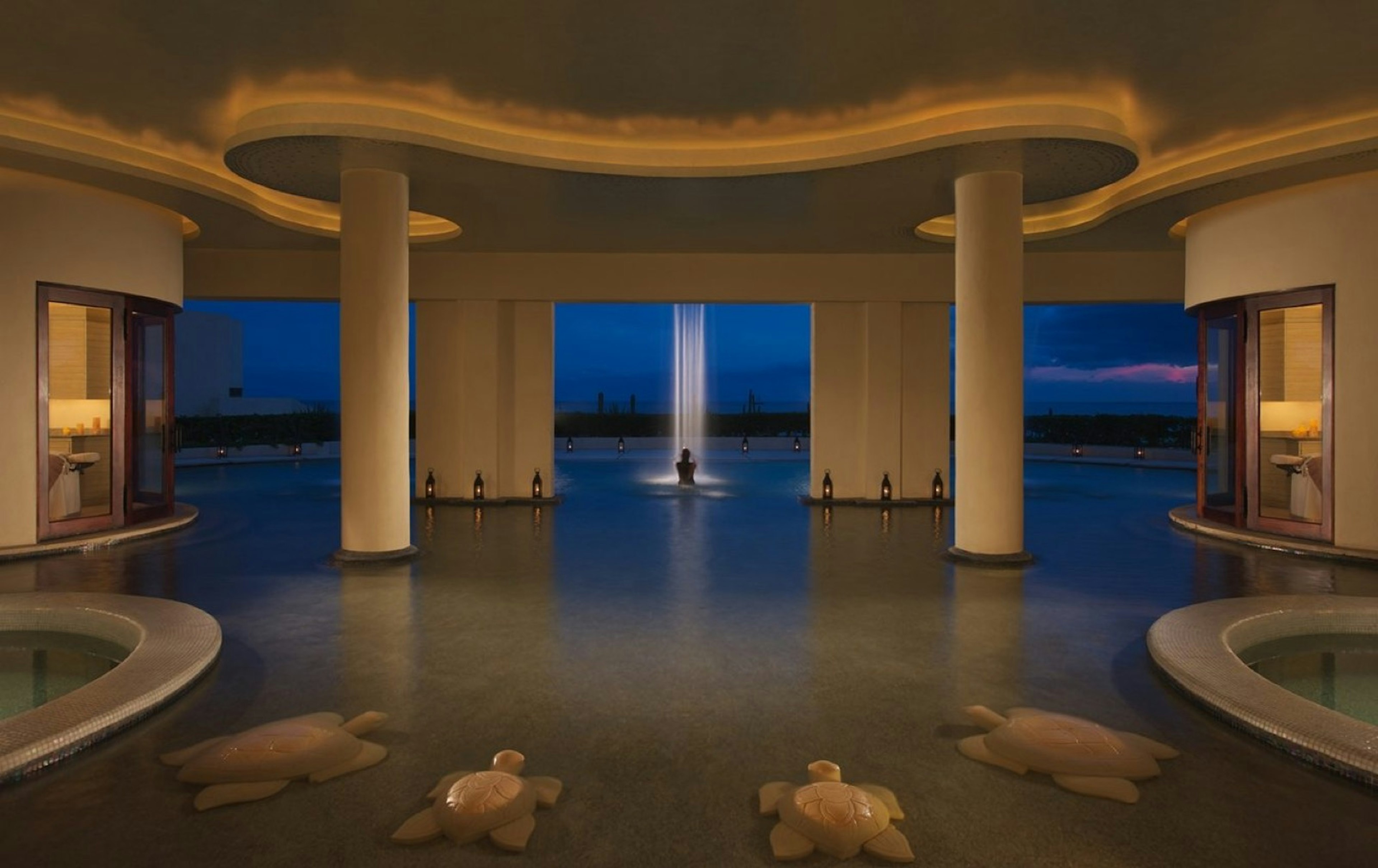 A woman stands beneath a waterfall in a lit pool at night