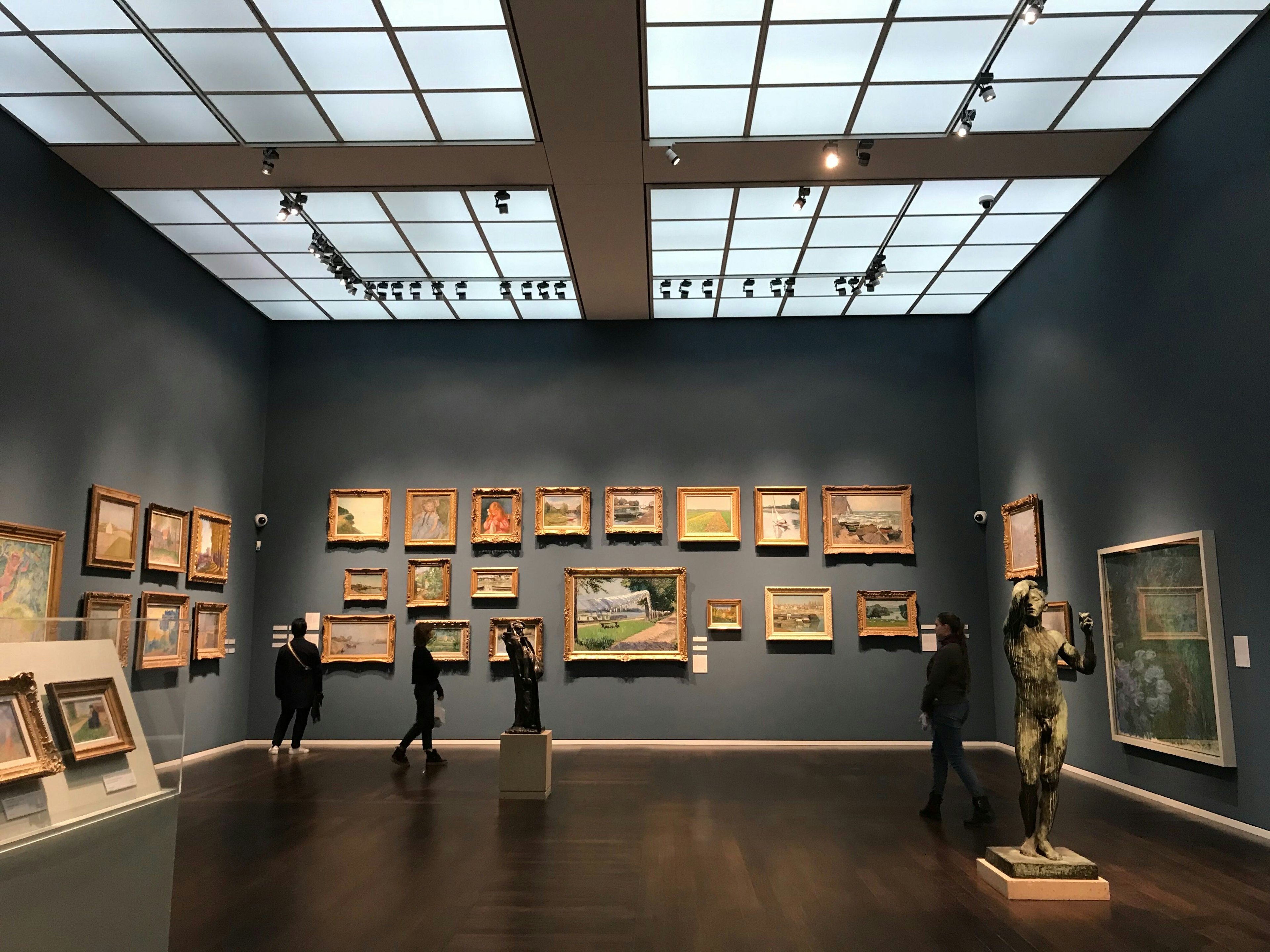 A series of colourful paintings with gold frames hang on a grey wall of a museum. There is a sculpture of a man in the foreground