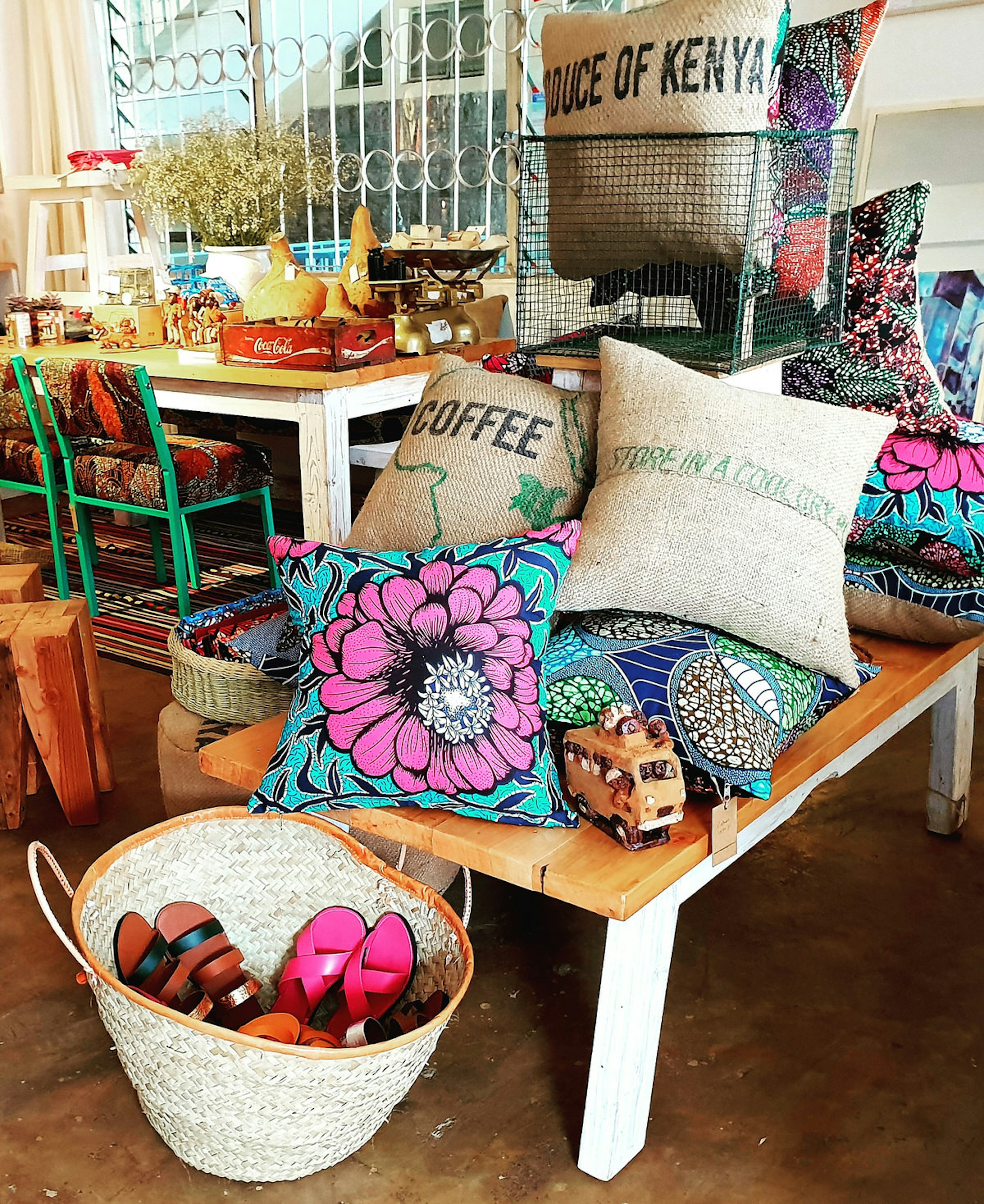 In the foreground sits a woven wicker baskey with brightly-coloured leather sandals. Immediately behind is a little wooden table with white legs that is piled high with colourful throw cushsions, as well as some made from hessian coffee sacks. A little toy truck is also on the table. Another fun place for Nairobi shopping © Clementine Logan / Lonely Planet