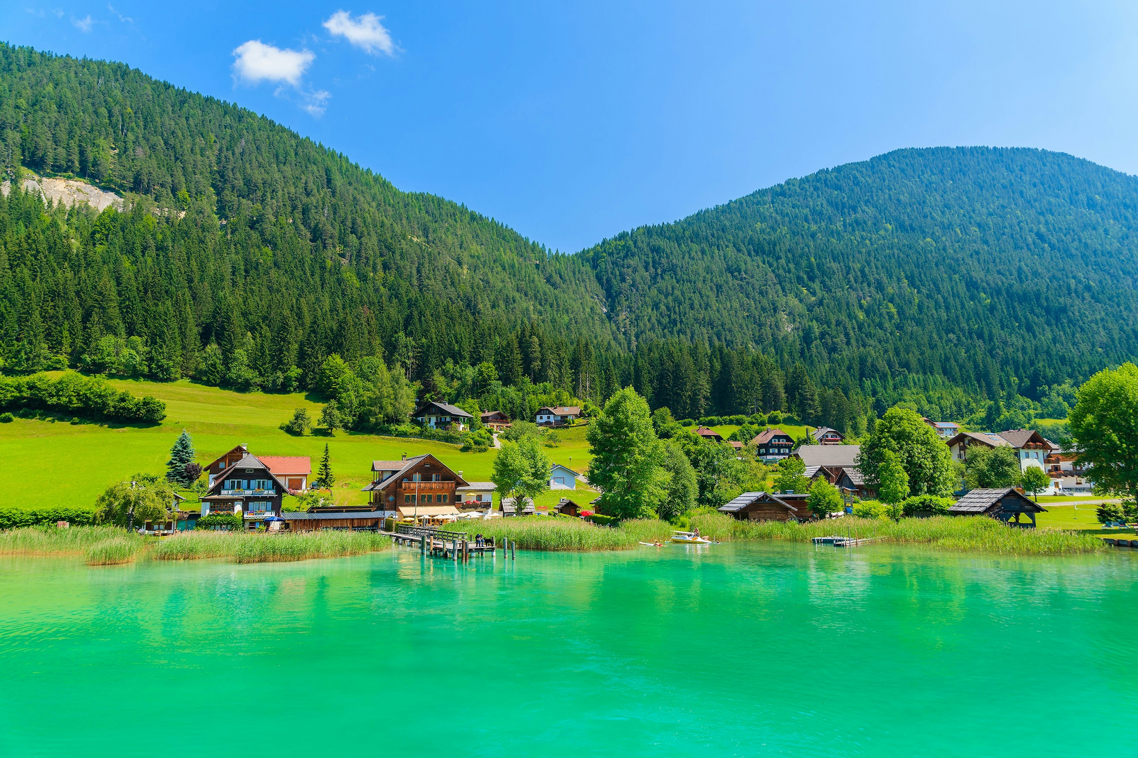 A bright green lake surrounded by hills covered in woodland. There are several chalet-style houses on the lakeside