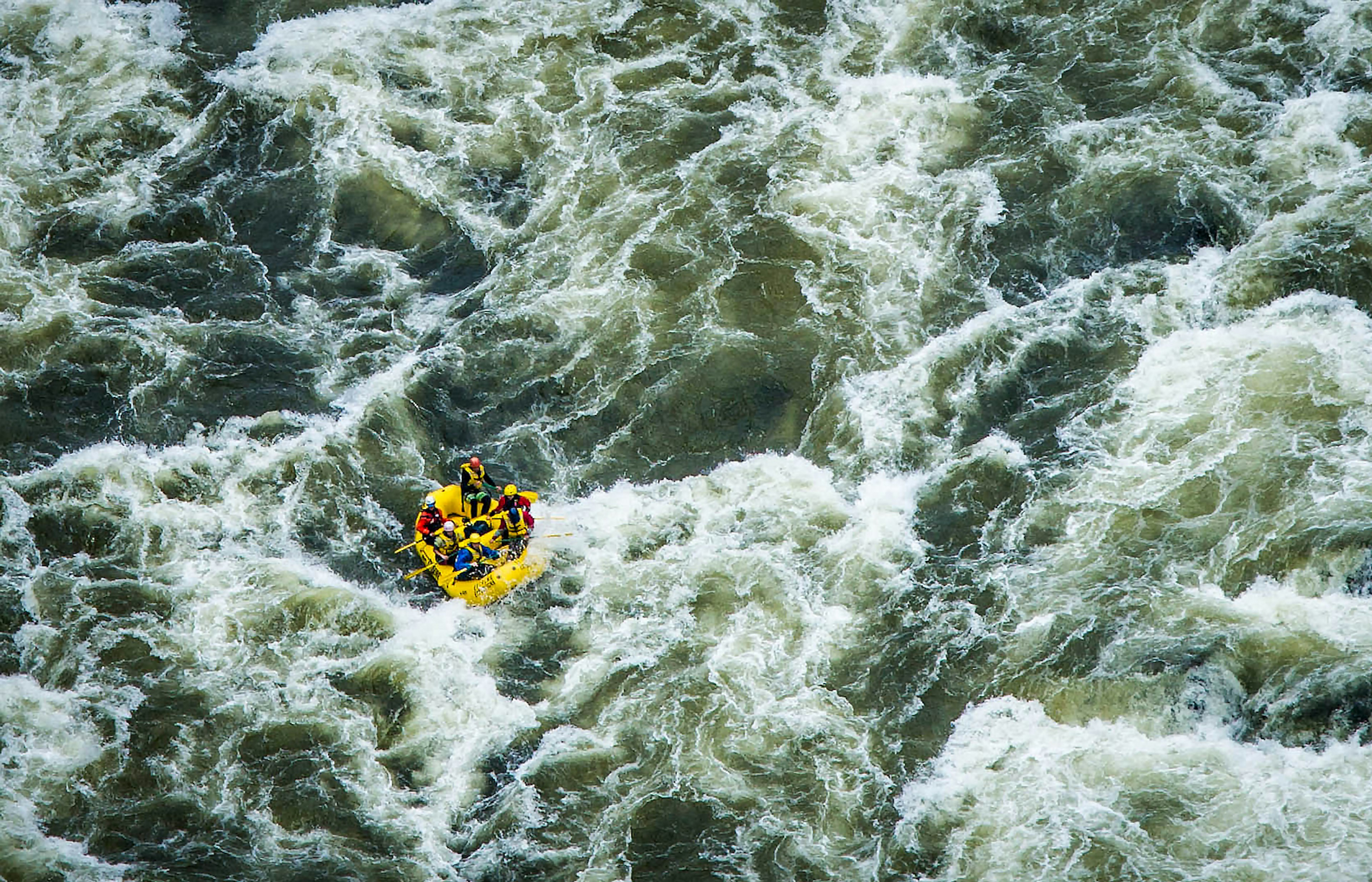 A yellow boat with six people aboard looks small from the aerial view of white water waves.