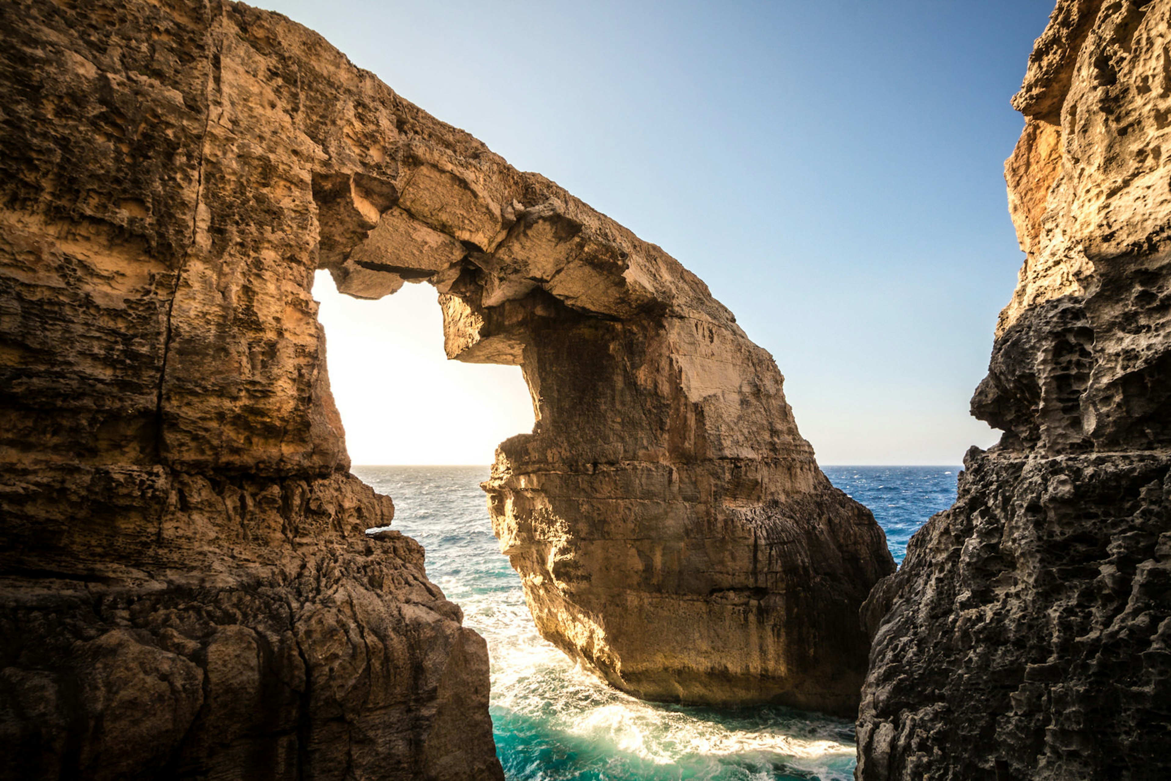 Sunlight bursts through a huge, angular rock arch on the coast of Goza at Wied il-Mielah. Wave surge between the two rock arms © Ramon Portelli / Getty Images