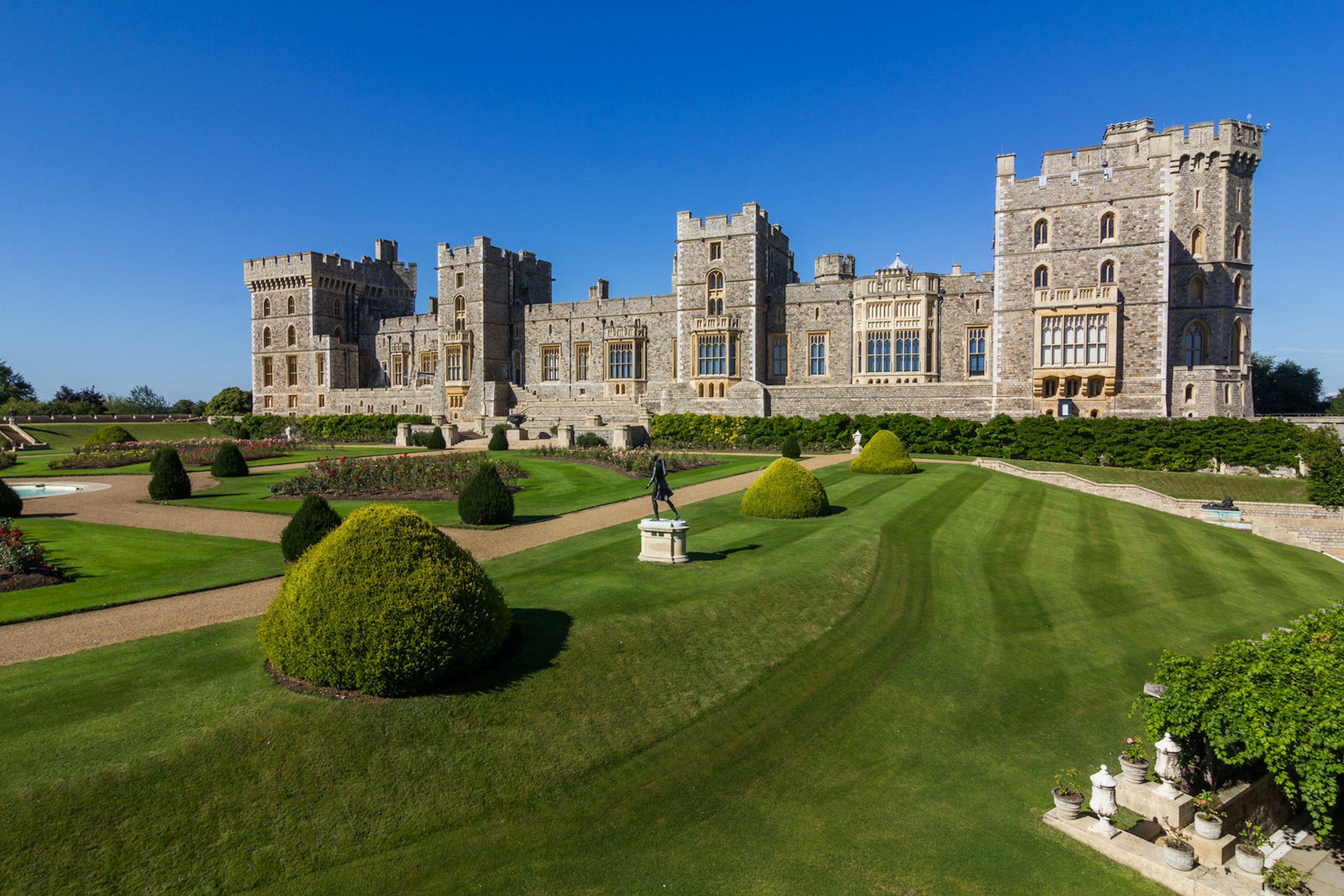 Windsor Castle's grey stone walls and castellated towers rise into a clear blue sky, with the extensive manicured lawn, hedges and shrubs laid out in the foreground; the castle is a royal wedding site © Kanuman / Shutterstock