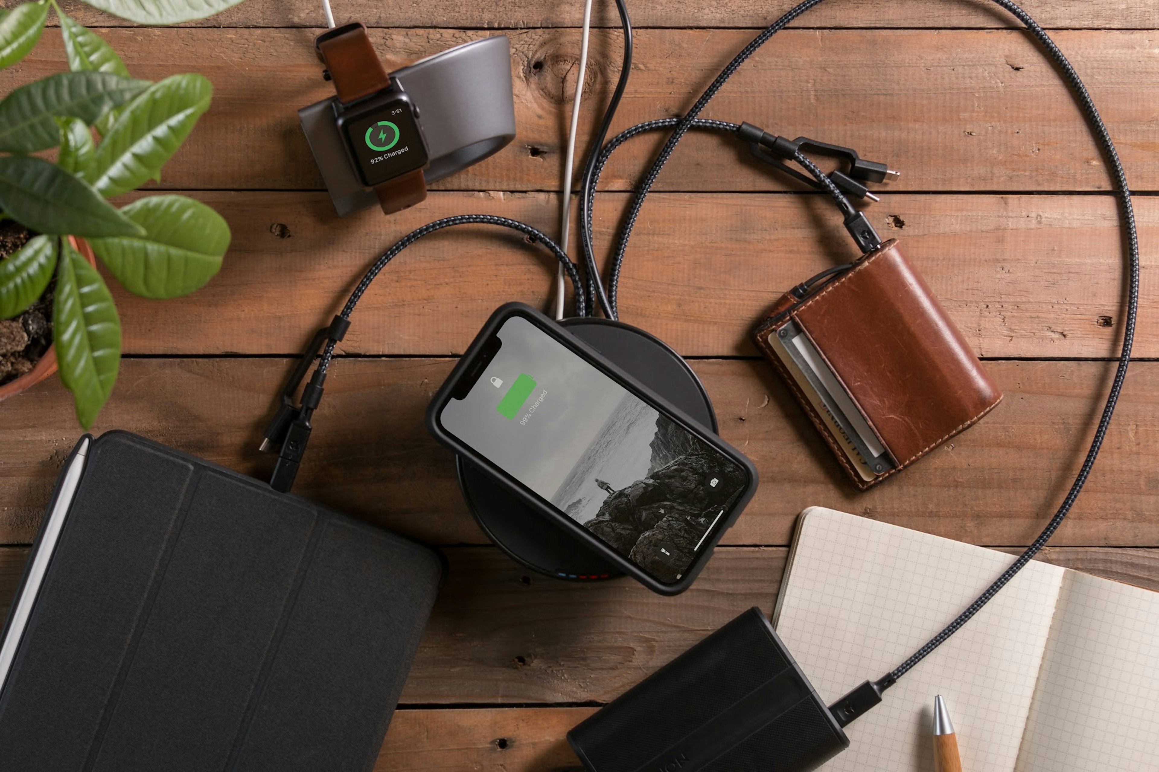 A table made of wide, wooden planks in a coffee-color holds a small plant to the left, a black iPad or similar tablet, a locked iPhone or similar smart phone, a black portable battery, a notebook with cream-colored grid-lined pages, and a warm brown wallet that is being use to charge all the devices from a black charging hub