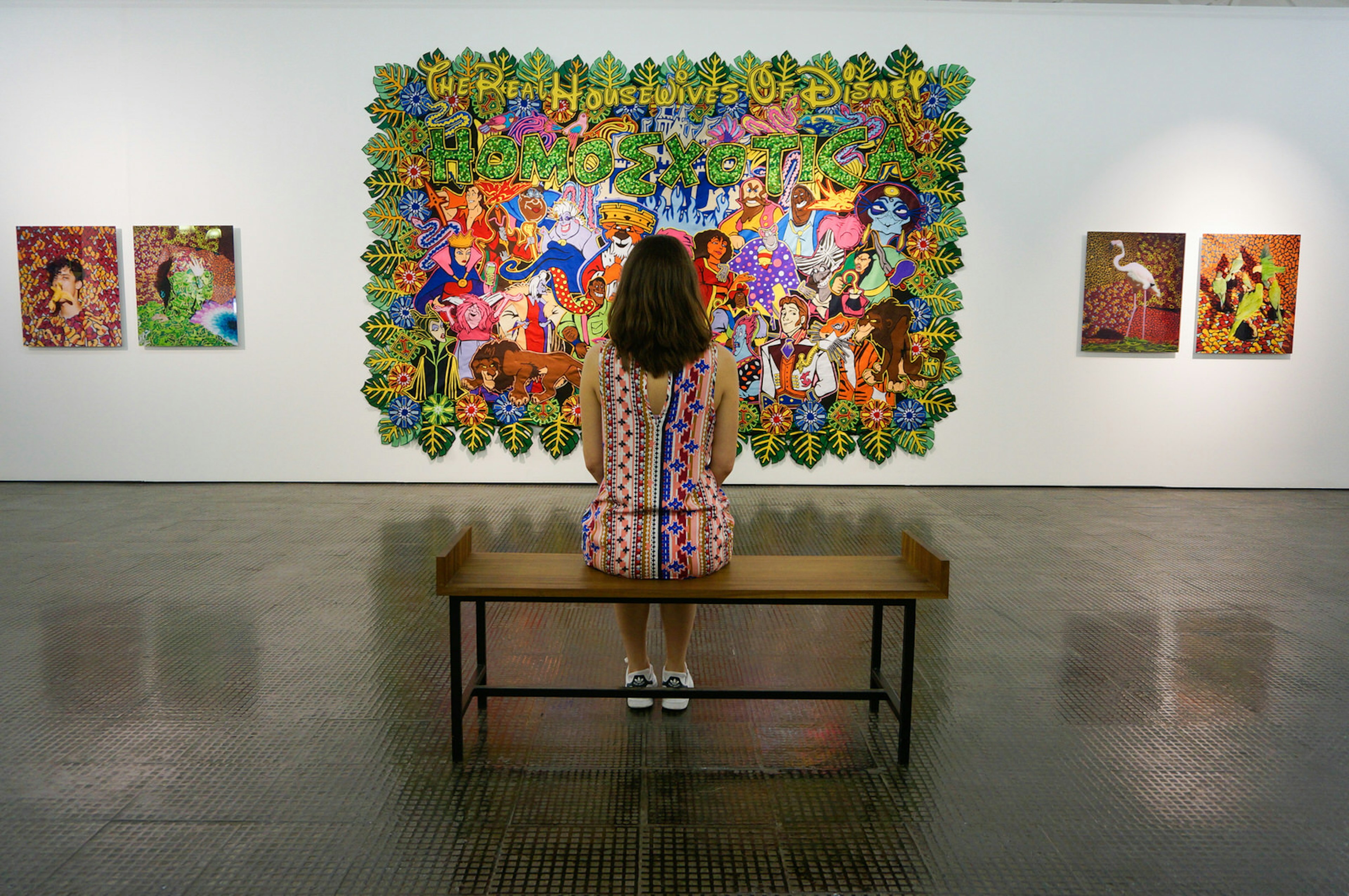 A woman sits in the middle of a simple bench with her back to the camera. In front of her are a number of artworks hanging on the wall, one featuring 'The Housewives of Disney', a colourful-cartoon painting © Monica Suma / ϰϲʿ¼