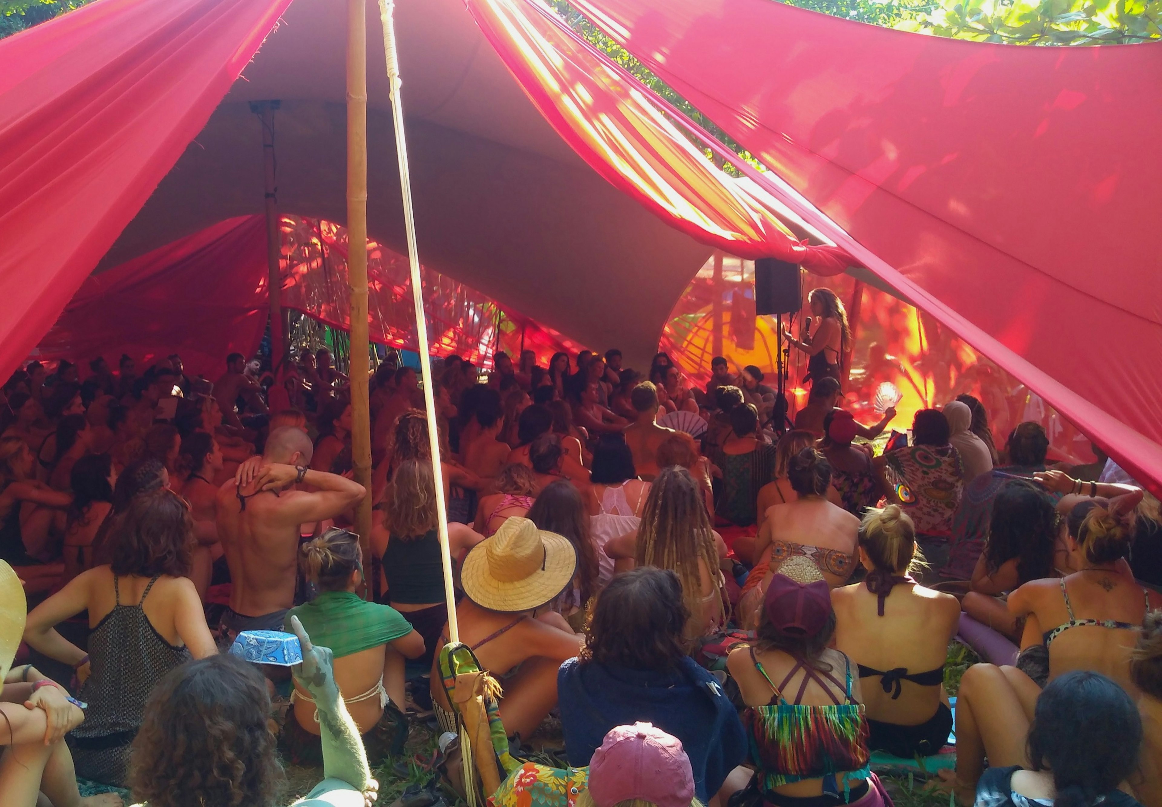 Dozens of women sit in a shady cloth tent as they listen to a speaker talking with a microphone