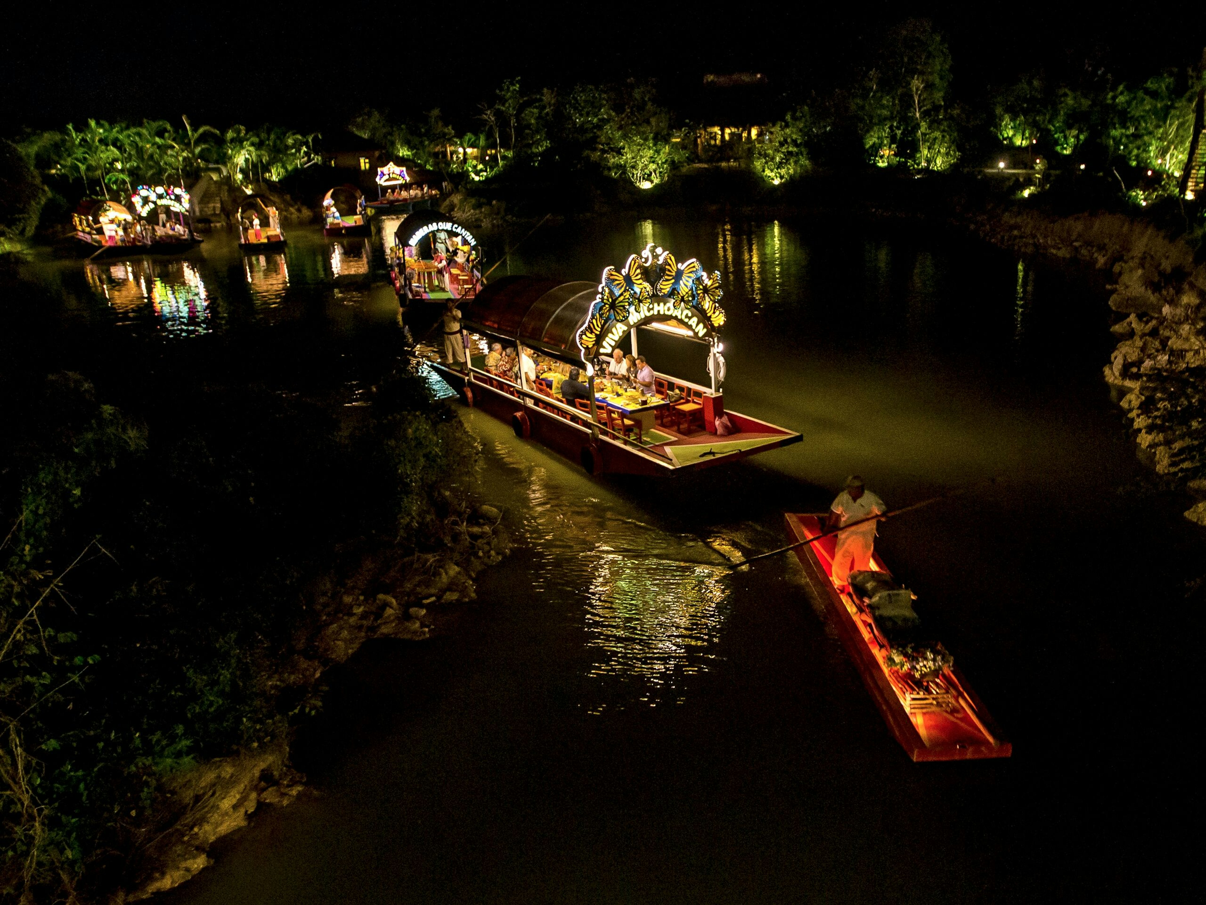 A series of gondola-like boats glide across the Yucatan Canal at night. All the boats have brightly lit signs at the front of the boat.