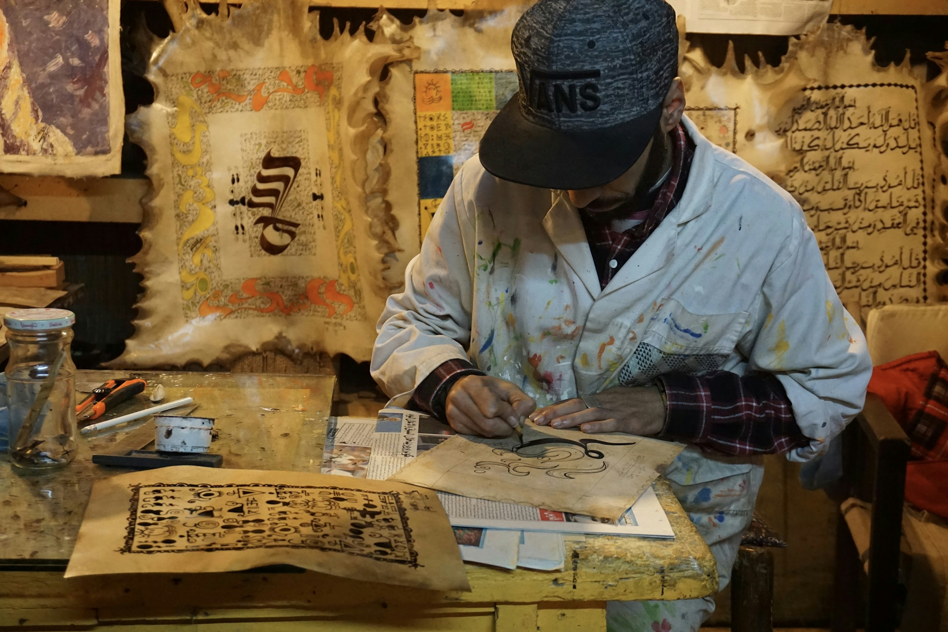 A local artist painting traditional Berber symbols onto canvas in his workshop; many of his works crowd the table he is working on and hang from the wall behind him.