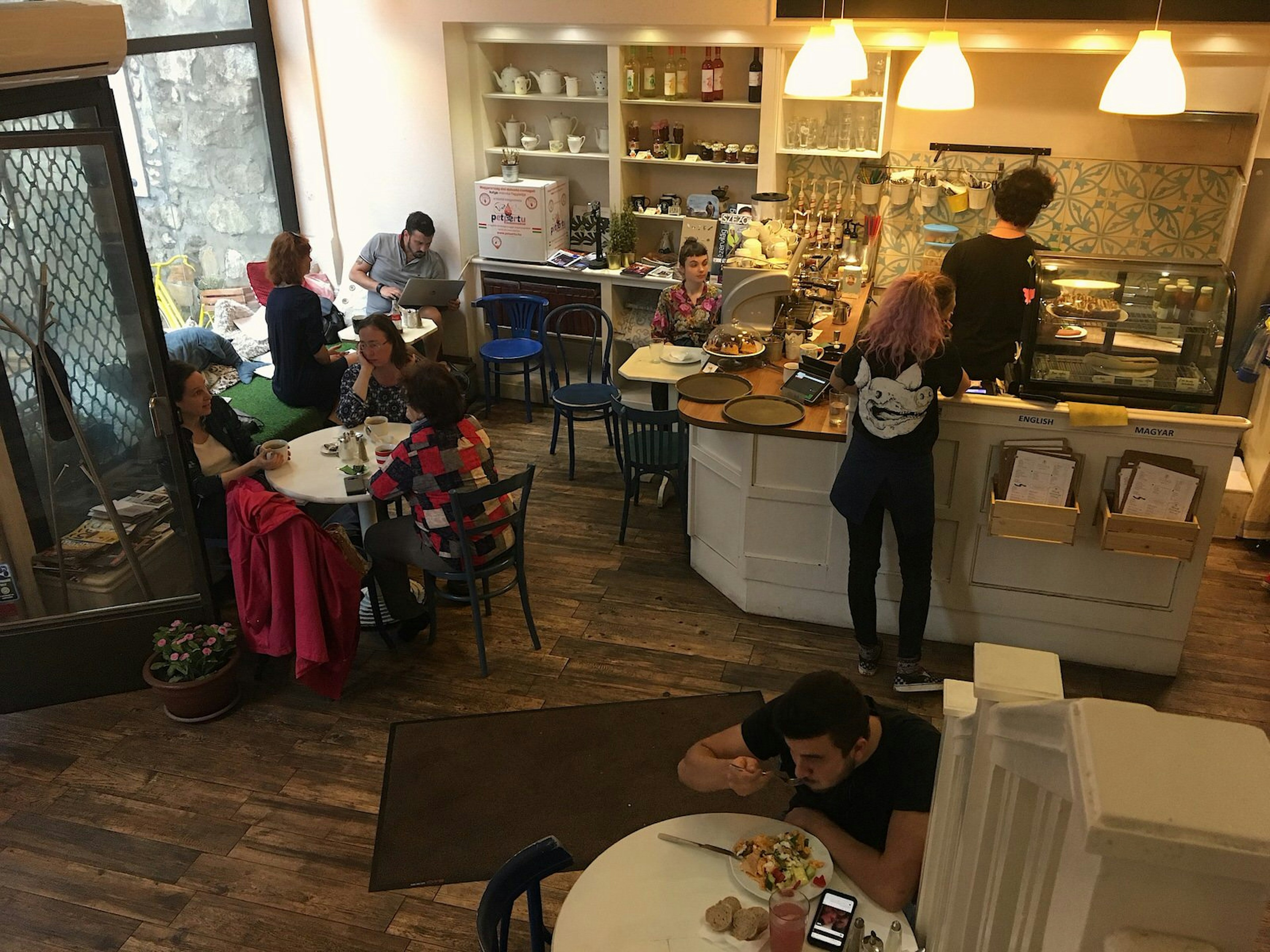 Diners fill tables near floor-to-ceiling windows while a customer orders at the counter; a man in the foreground has fork in mouth and a plate full of colourful food